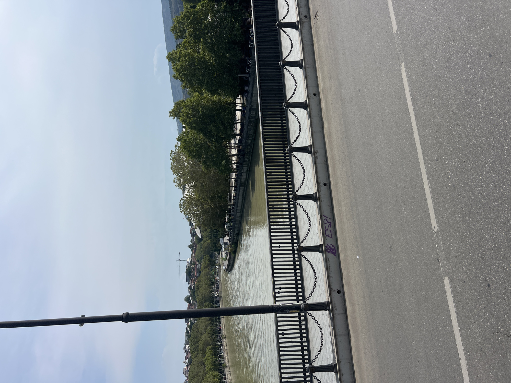 This is an eye-level, medium shot capturing a view across a bridge in Tbilisi, Georgia. The foreground consists of an asphalt road with a white lane marking. Beyond the road is the pedestrian walkway of the bridge, separated from the walkway by a metal railing with a decorative chain suspended between posts. This railing has graffiti on its base, including purple scrawled text. The background features a wide, murky river, with a lush green riverbank lined with trees and buildings. In the distance, a construction crane is visible amongst the buildings. The sky is partly cloudy. There are no visible people or animals in the frame.