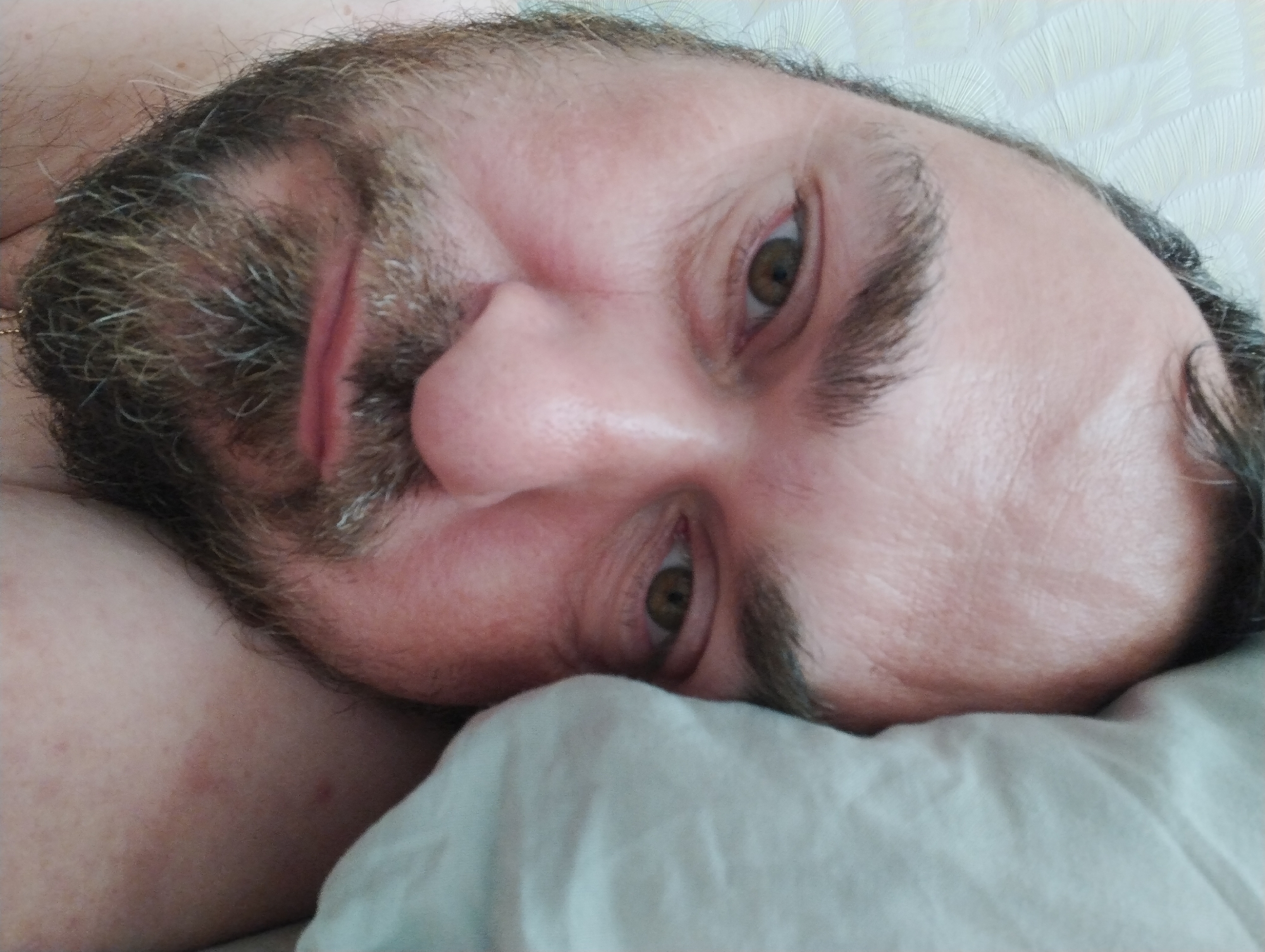 An extreme close-up shot shows a Caucasian man's face, lying on his side, with his head tilted. The man has brown hair and a beard with some gray strands. His eyes are open, and he appears to be looking towards the camera. The lighting is soft, and the background is a textured white surface, possibly a bedsheet. There are no other people or objects visible in the image. The image does not contain any contextual information about the city of Konstantinovo, Russia.
