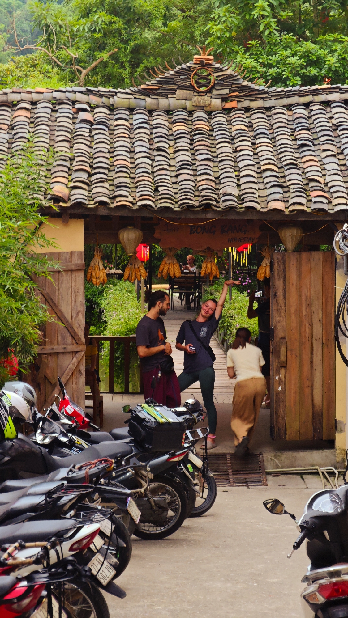 A full shot captures the entrance to Dong Bang Homestay in Yên Minh Commune, Vietnam. The entrance is distinguished by a tiled roof, wooden door, and signs. A group of people are present in front of the homestay. A man and woman appear to be tourists, engaged in conversation. A woman in a tan outfit walks through the doorway into the homestay. Several motorbikes are parked in the foreground. Decorations such as lanterns and what appear to be corn cobs adorn the entrance. Green foliage is visible above the entrance, and a bridge or walkway extends into the background.