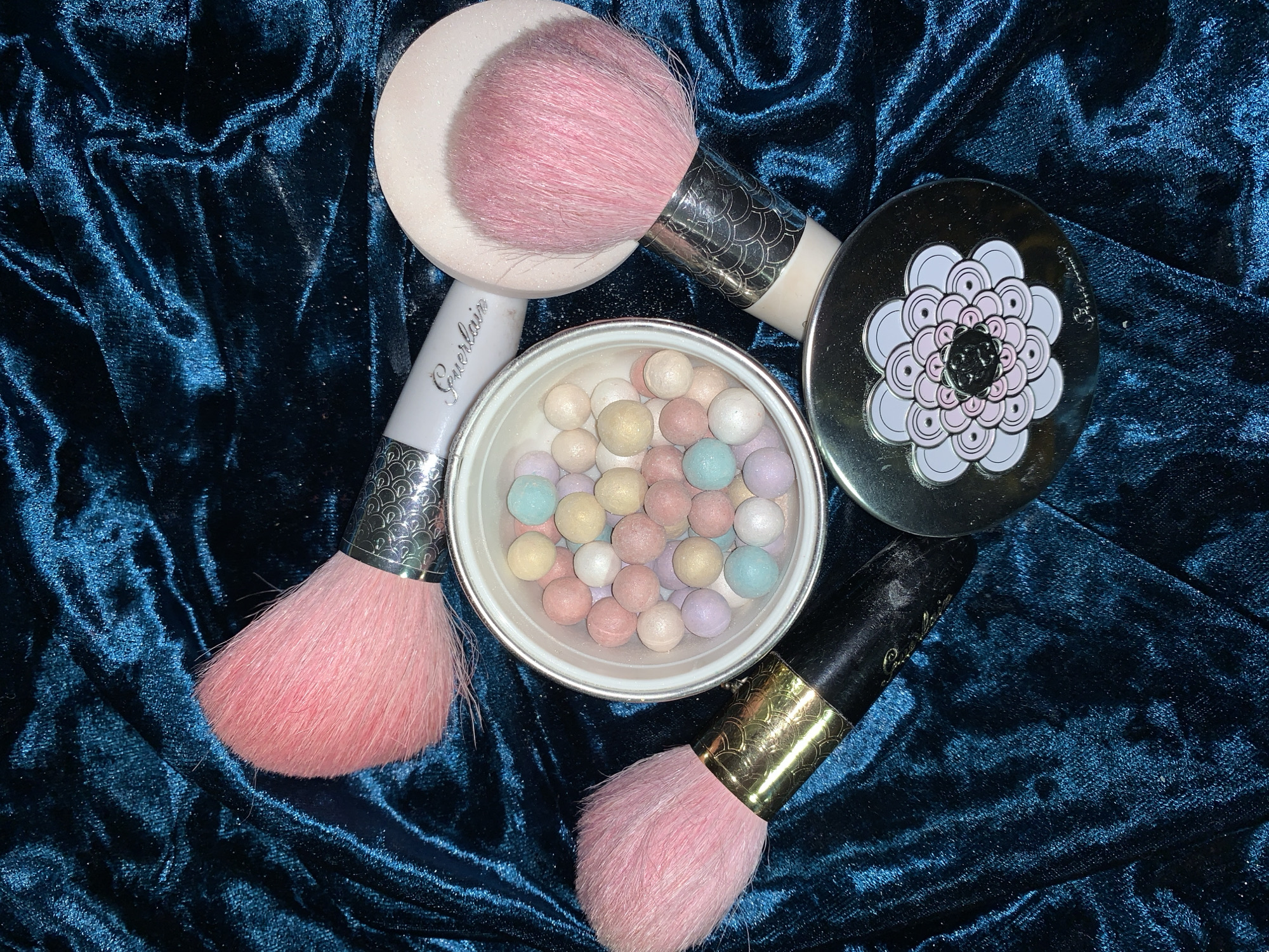 The image shows a close-up of several makeup items arranged on a textured dark blue surface. A container of Guerlain Meteorites pearls is centered, surrounded by three pink makeup brushes. The lid of the Meteorites container is visible on the upper right. The background is a crumpled, possibly velvet or similar plush fabric, creating highlights and shadows. No people are visible in the image.