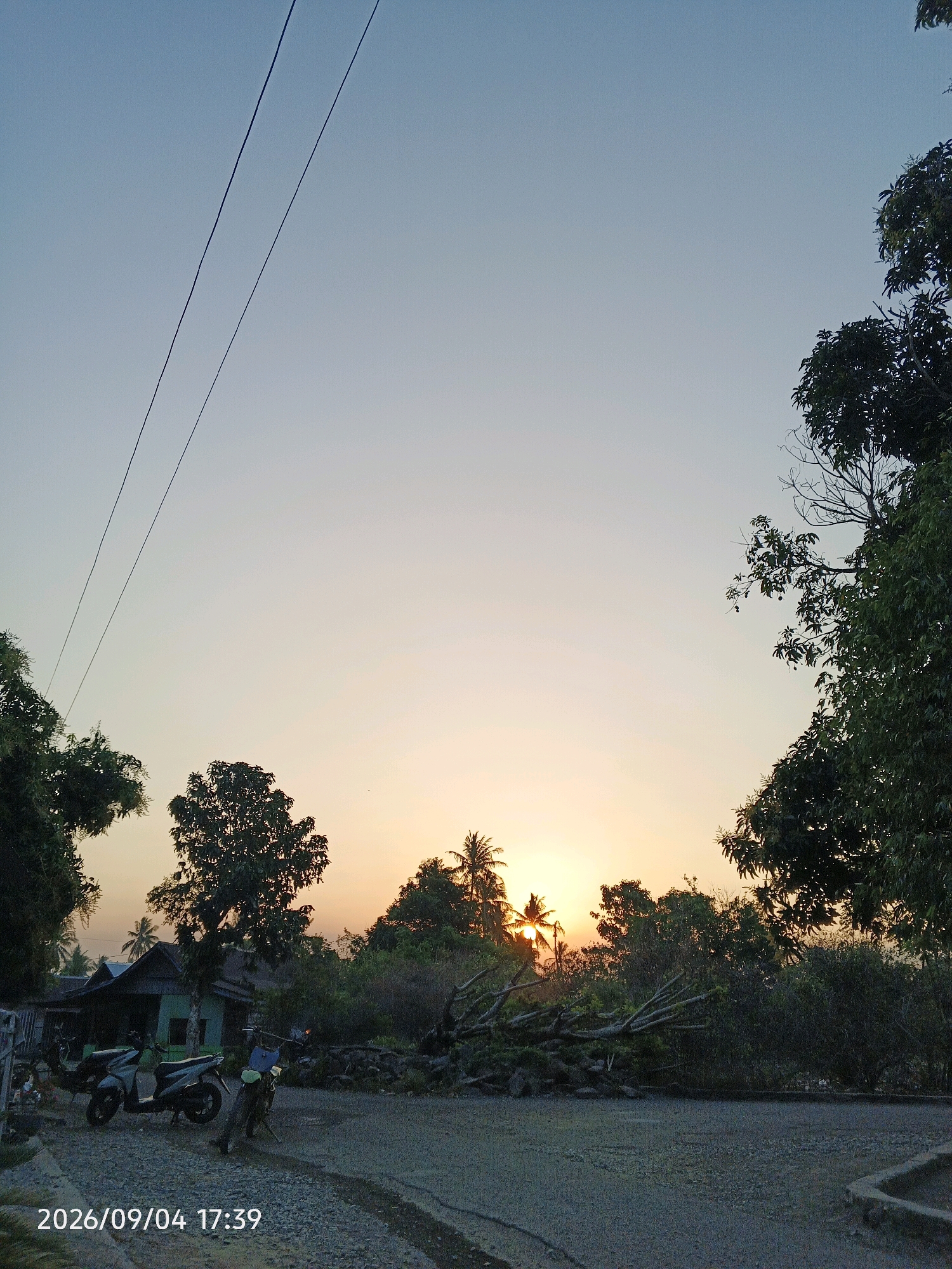 This is a photograph taken at sunset in Bulu Cenrana, Indonesia. The image captures a rural scene with a dirt road in the foreground. Two motorcycles are parked on the left side of the road. Behind the motorcycles, a small house with a dark roof and a light-colored wall is visible. A cluster of trees frames the sunset in the background, with the sun partially obscured by foliage. Power lines are visible in the upper left quadrant of the image, stretching across the sky. The sky transitions from a light blue at the top to a soft orange and yellow hue near the horizon, indicating the setting of the sun. The overall atmosphere is serene and natural.