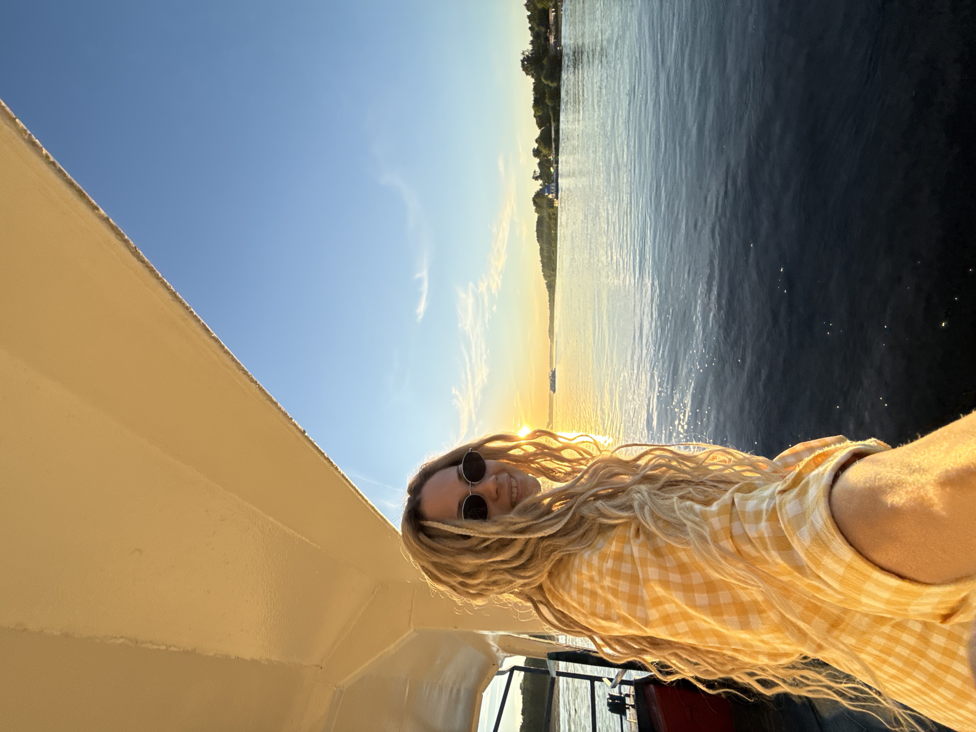 An eye-level medium shot taken on a boat in Заволжье, Russia, during sunset, captures a young woman with long blonde dreadlocks and dark oval sunglasses. She is positioned on the left side of the frame, smiling faintly towards the camera, her body angled towards the open water and the setting sun on the right. She wears a yellow and white gingham long-sleeved shirt. Her hair and the right side of her face are brightly illuminated by the golden light of the sun, which is visible as a distinct orb just above the distant horizon.
The setting sun casts a warm, golden glow across the expansive body of water, likely a river, that dominates the right and lower portions of the image. The water's surface exhibits subtle ripples and reflections. In the distance, a dark green, tree-lined shoreline extends horizontally, with a few light-colored buildings interspersed among the foliage. At least two other watercraft are visible further down the river. The sky transitions from a bright golden-orange near the horizon to a clear, pale blue at the top right, with minimal cloud cover. The upper left quadrant of the image shows the underside of a boat's light-colored deck or canopy structure, with a white metal railing or support visible behind the woman on the left.