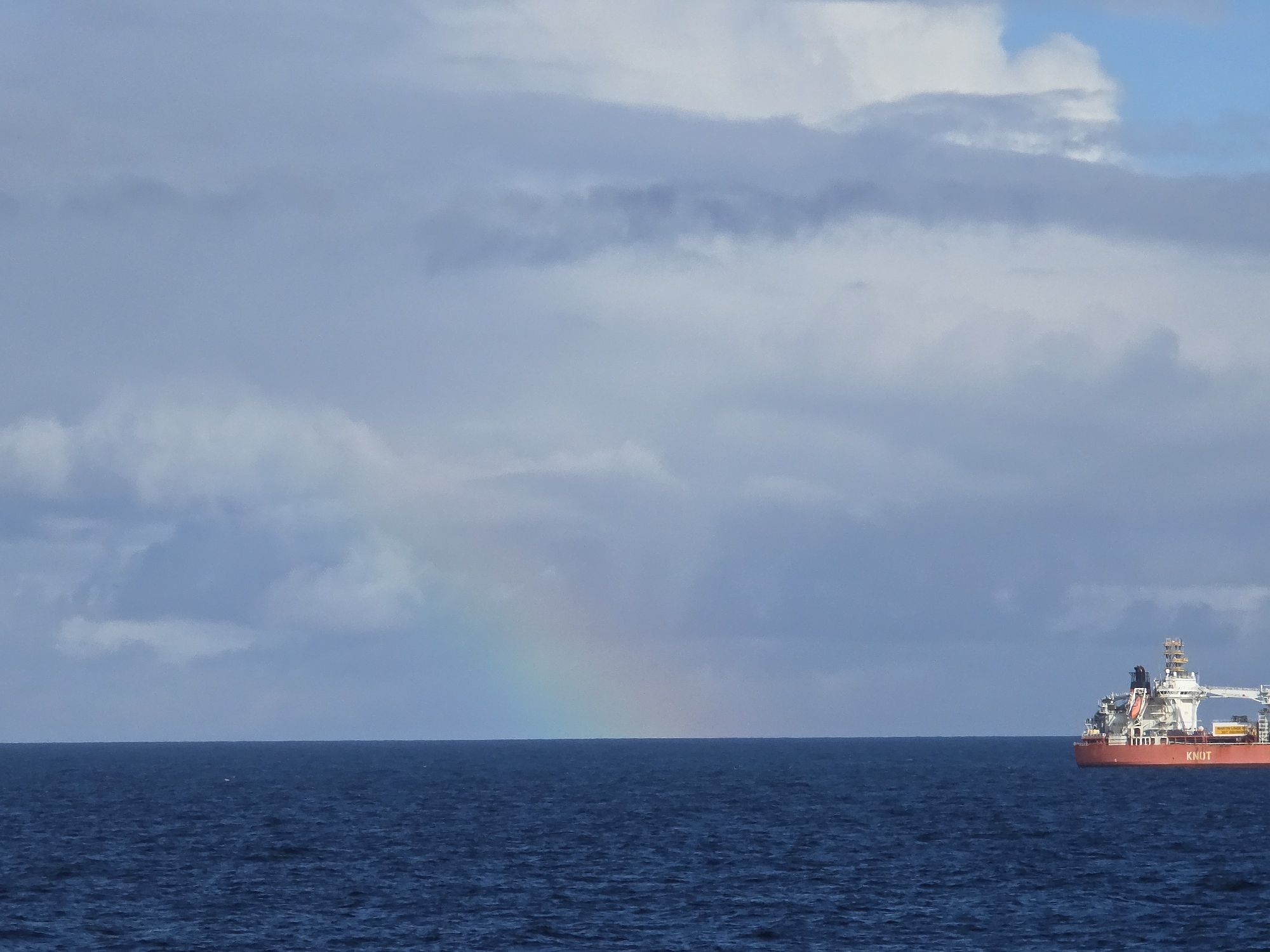 The image depicts a seascape under a cloudy sky, where a faint rainbow arches over the horizon. A red and white ship is visible on the right side of the frame. The location is Unknown, Unknown.
