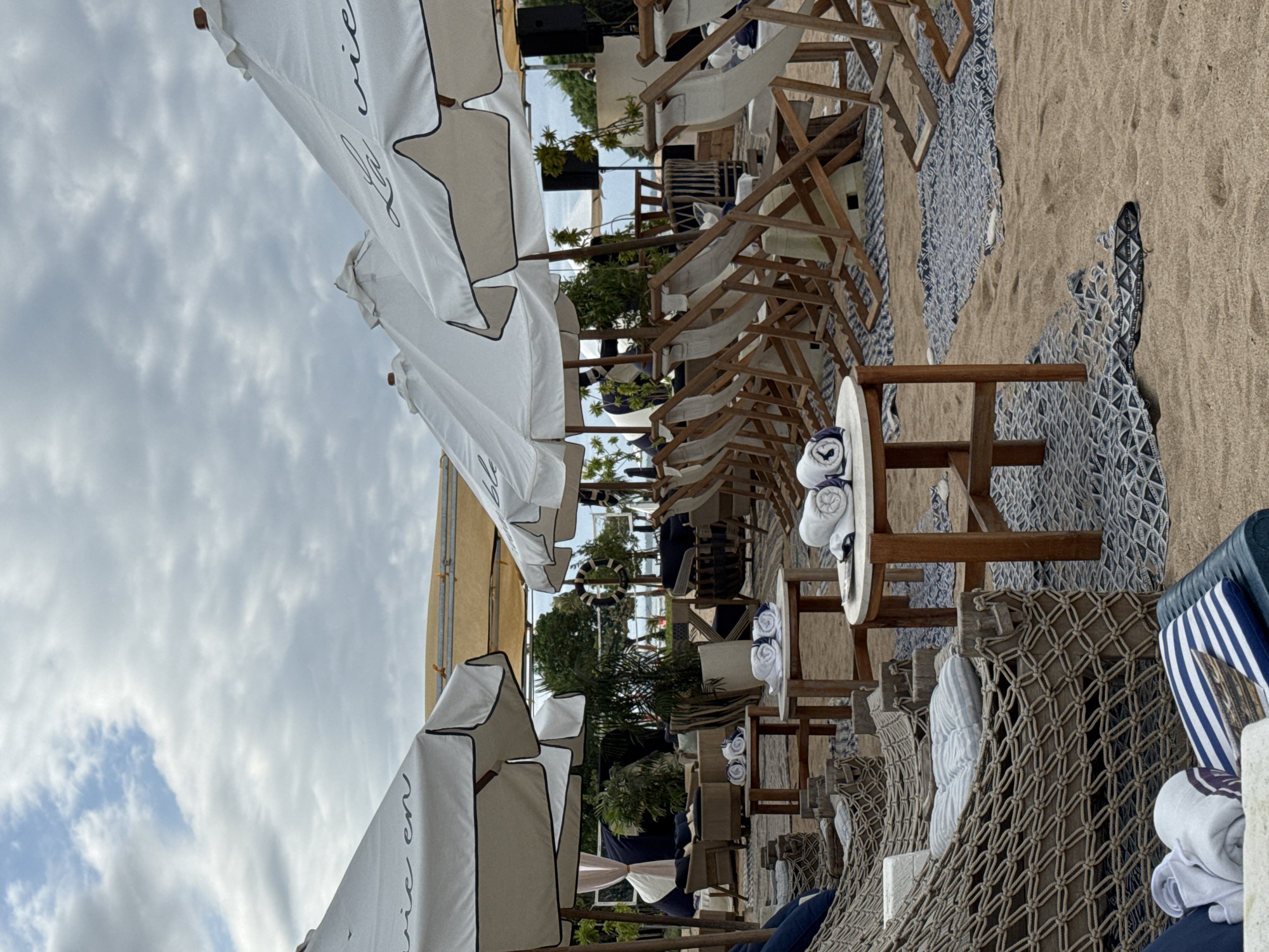 The photograph depicts a beach scene in Kuta Selatan, Indonesia. Rows of beach chairs with wooden frames and white fabric are arranged on the sand. Beside each chair is a small round table with neatly rolled white towels on top. White beach umbrellas with black trim are positioned over the chairs, partially obscuring the sky, which is filled with gray clouds. The ground is covered in patterned cloths beneath the chairs. In the background, lush green foliage and other beachside structures can be seen.