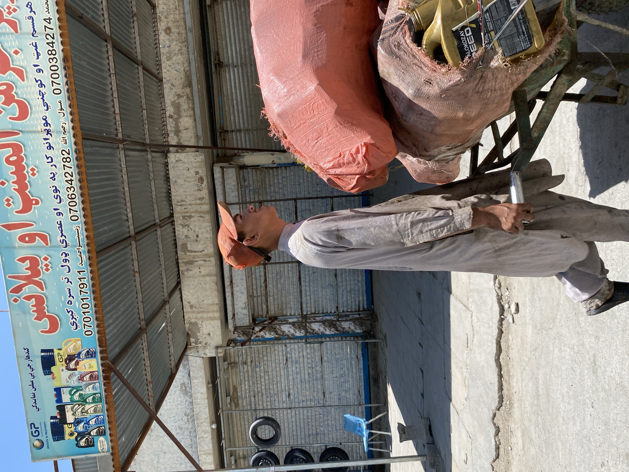 In Kandahar, Afghanistan, a young man wearing a light-colored shalwar kameez and an orange baseball cap walks past a cart loaded with sacks and yellow oil containers. The cart is positioned on a paved area, with a cracked and uneven surface. Behind him, a store with a large blue and red sign displays various oil cans and includes text in Pashto and numbers: "0700384274" and "0706342782". The store appears to be a retail establishment, possibly an auto parts or oil supplier, with metal roll-up doors closed behind the sign. A few tires are stacked on a rack to the left, and a blue plastic chair sits nearby. The overall environment suggests a commercial area within the city, under a bright, sunny sky.