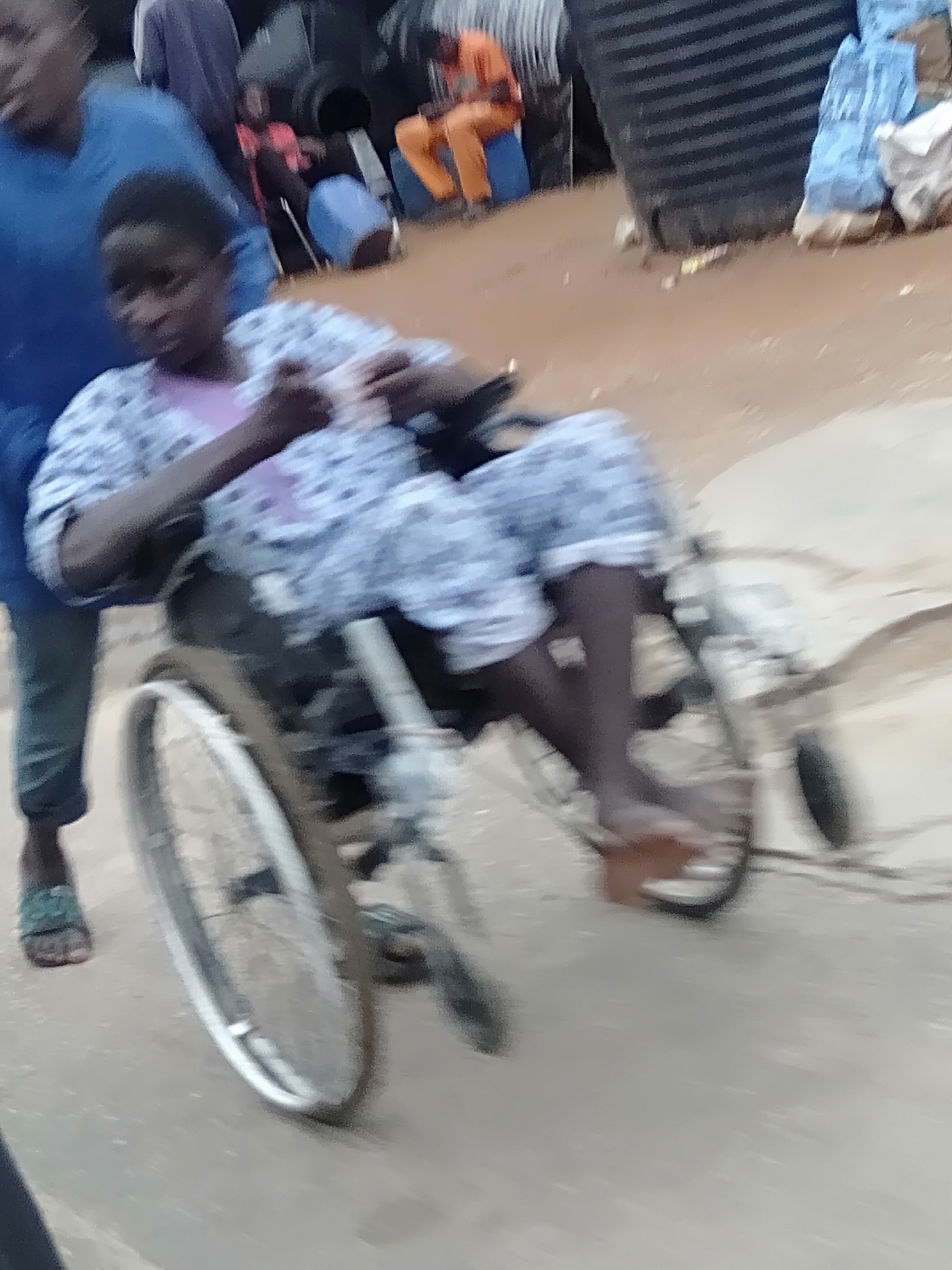 A medium outdoor shot in Unknown, Unknown, focuses on a person in a wheelchair being pushed by another. The person in the wheelchair is wearing a light-patterned pajama set with a light-colored shirt underneath. They are barefoot and appear to be holding something in their hands. The person pushing the wheelchair is wearing a blue top and blue pants with green sandals. In the background, there are blurred figures, large black containers, and various other objects. The ground appears to be a combination of dirt and pavement. The overall impression is one of a street scene in an impoverished area.