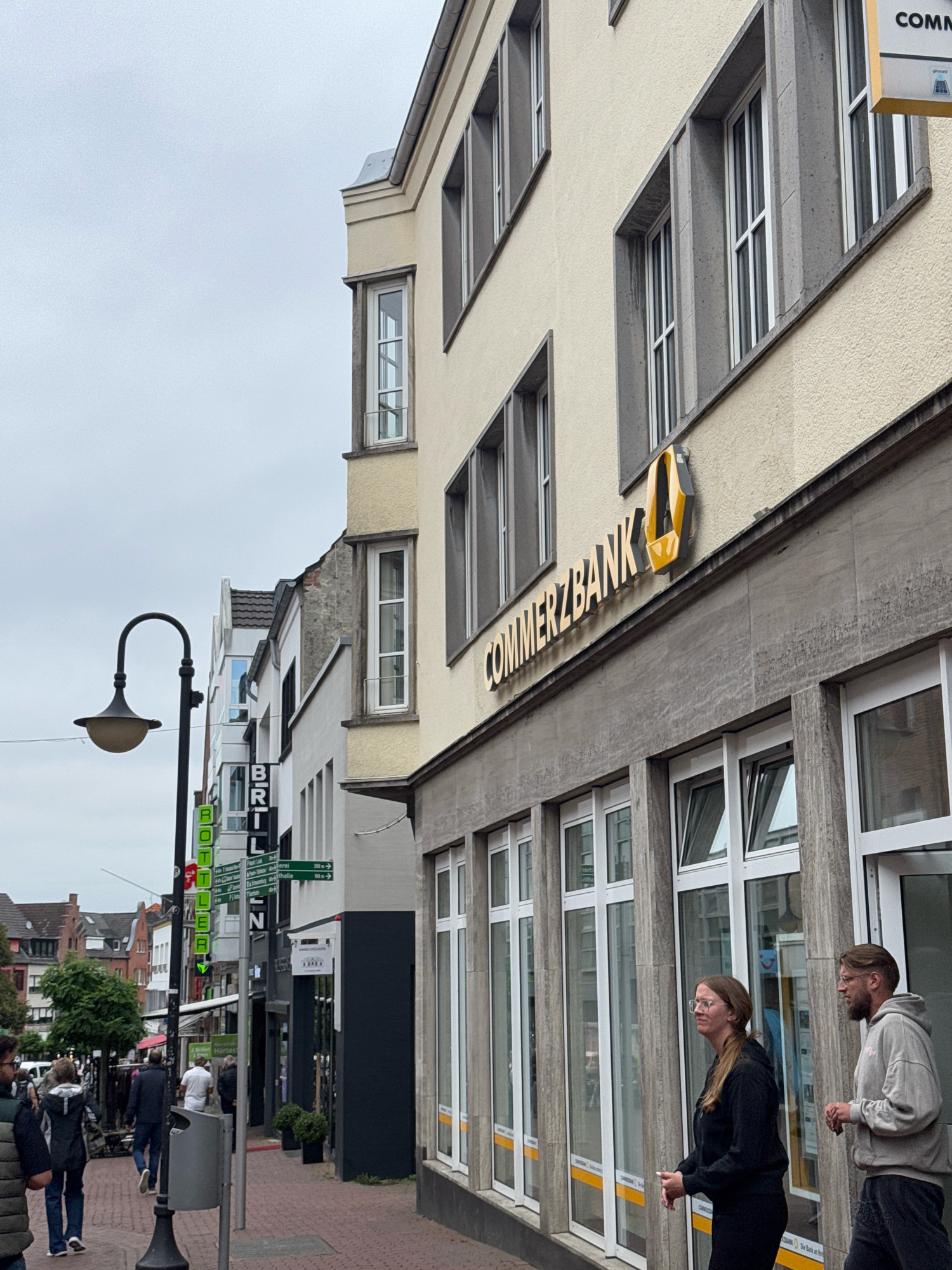 The image shows a street scene in Cleves, Germany. A modern building with a light yellow facade and concrete accents dominates the right side of the frame. The building features large, multi-paned windows. A sign for "COMMERZBANK" with a distinctive yellow logo is prominently displayed on the facade. Two people, a woman wearing glasses and a dark hooded sweatshirt, and a man wearing glasses and a light gray hooded sweatshirt, are walking past the bank.
To the left of the Commerzbank building, there is a row of older, narrower buildings with varying architectural styles. A black lamppost with a curved arm and a globe-shaped light fixture stands in the foreground. Several green and white signs are attached to poles, including one with the word "BRILL" and others with directional arrows indicating distances to locations like "Kneipp". Pedestrians are visible further down the street, and some shops appear to be open. The sky is overcast, suggesting a cloudy day. The street is paved with brickwork.