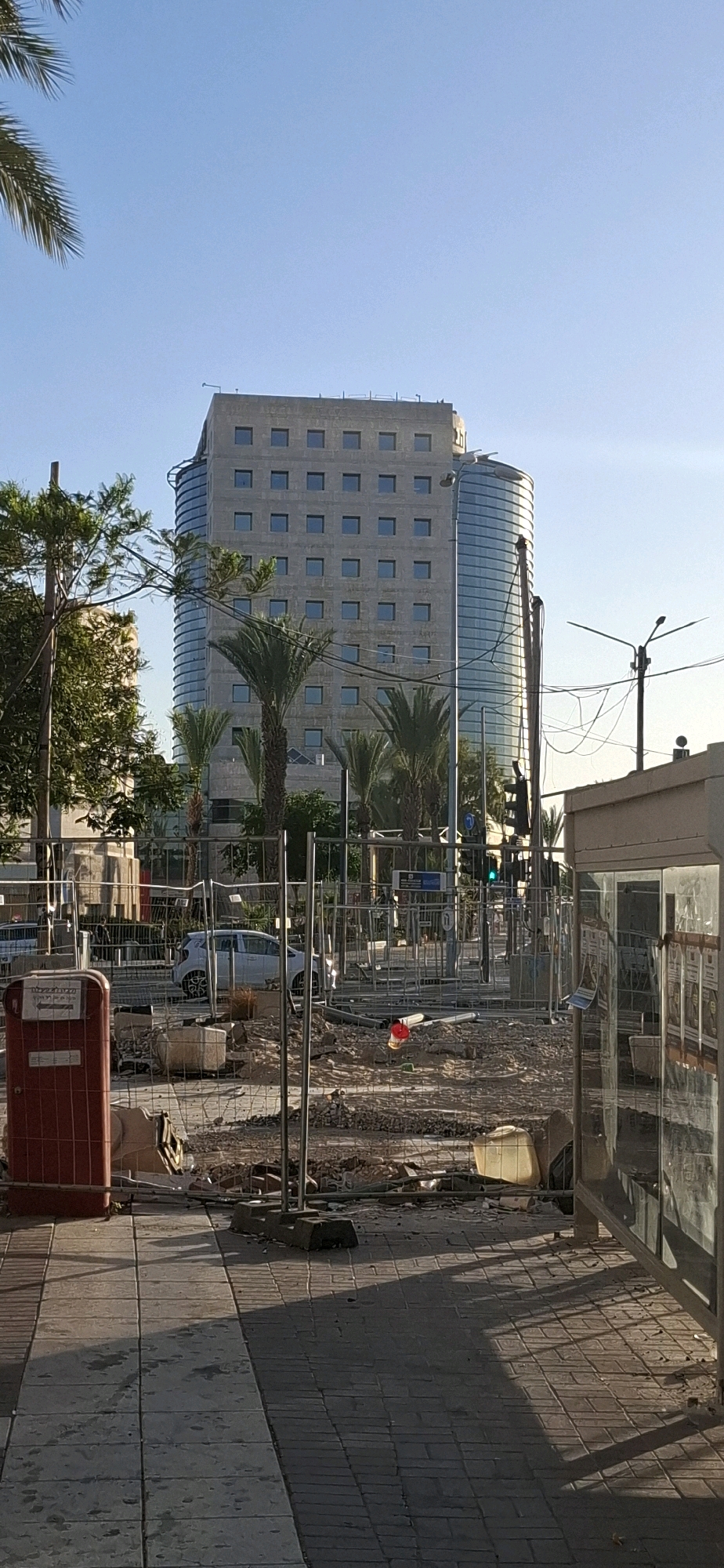 This is a medium-shot, vertical, outdoor photograph taken on a clear day with a bright blue sky. In the background, a tall, modern office building with a curved glass facade on the right side and a rectilinear concrete facade on the left side dominates the upper portion of the image. Rows of square windows are visible on the concrete section. In the midground, palm trees are sparsely planted in front of the building. A white car is parked behind a chain-link fence. The foreground shows a construction site with debris, sand, and concrete barriers. A red trash receptacle is on the left, and a bus shelter with glass panels is on the right. The ground is paved with interlocking stones, with shadows cast by the fence and the bus shelter. This scene is located on a street in Be'er Sheva, Israel, likely a commercial or downtown area. There are no people visible in the frame.