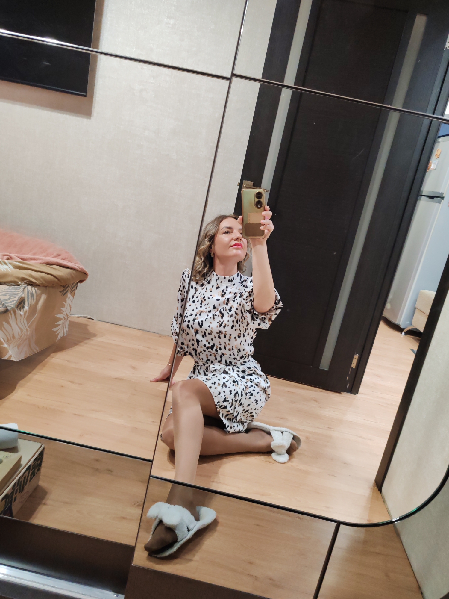 A middle-aged woman is reflected in a mirror taking a selfie. She is sitting on a wooden floor wearing a patterned dress and slippers. Behind her in the reflection is a bed, a dark-colored door, and a refrigerator. The location is likely indoors, possibly an apartment. There is no verifiable evidence to indicate that the location is Petropavlovsk-Kamchatsky, Russia.