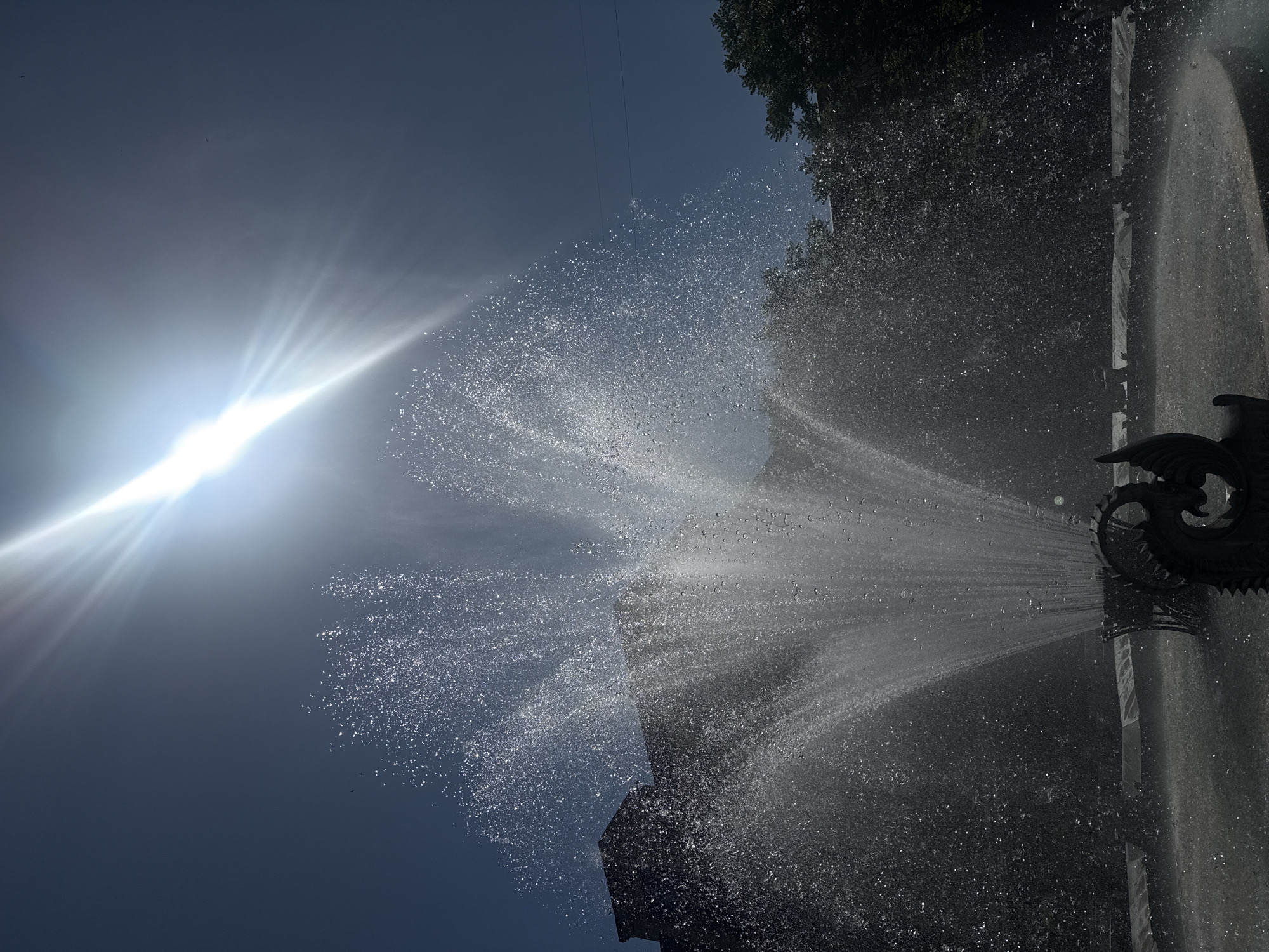 The photo depicts a fountain in Yerevan, Armenia, spraying water upwards against a bright, sunny sky. The fountain is constructed with ornate, cast-iron elements and the water spray is voluminous, catching the sunlight and creating a sparkling effect. Trees are visible on the right side of the image, and building silhouettes appear near the base of the spray. A wire runs along the top center of the image.