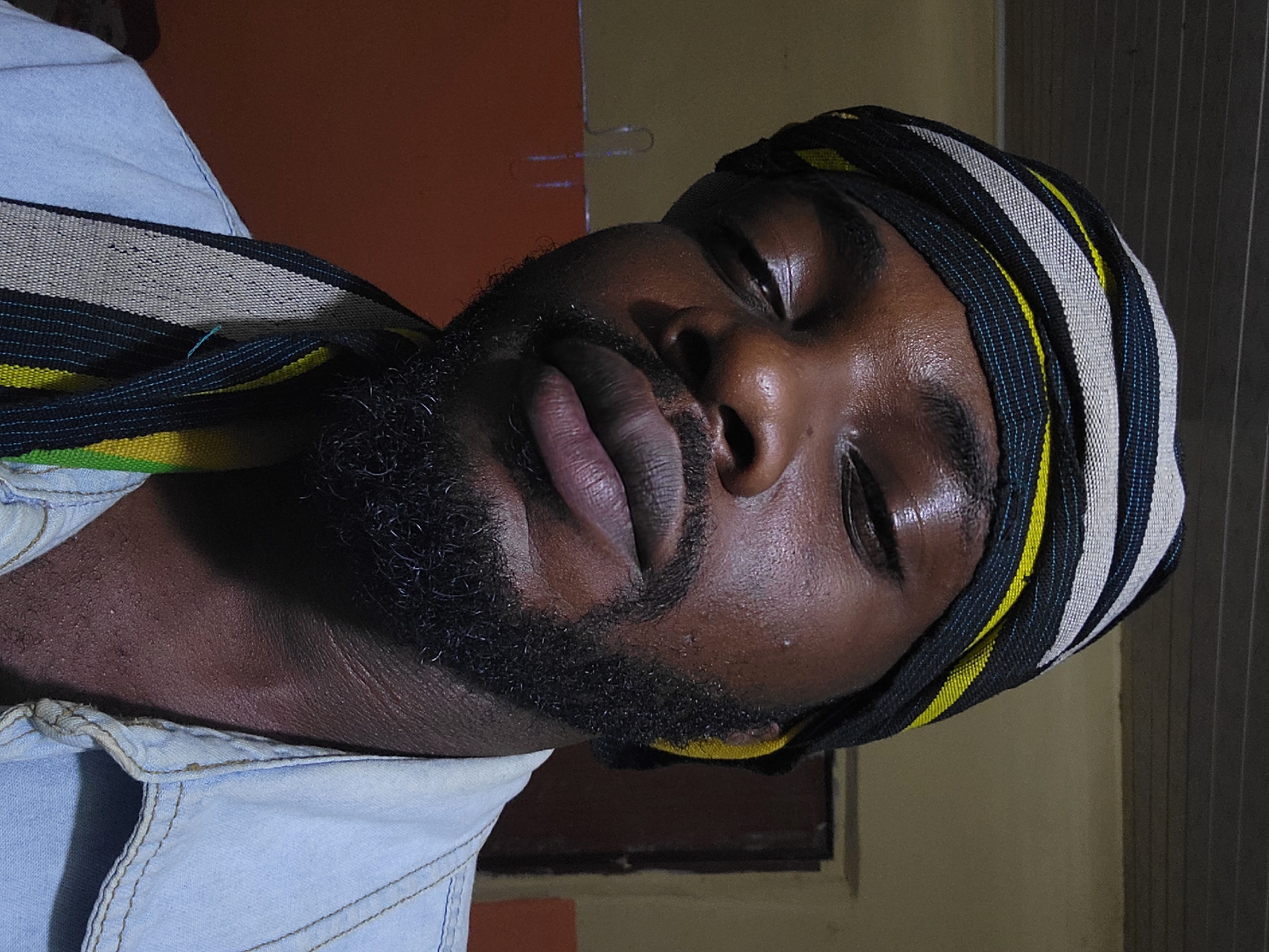 A close-up, portrait-oriented shot depicts a dark-skinned man, his eyes closed as if sleeping. He wears a head wrap with alternating black, white, and yellow stripes. A dark beard frames his face. He wears a light blue, denim-like shirt. The background shows an interior with walls of orange and beige, and corrugated metal. The city of Jos, Nigeria cannot be verified from this image.
