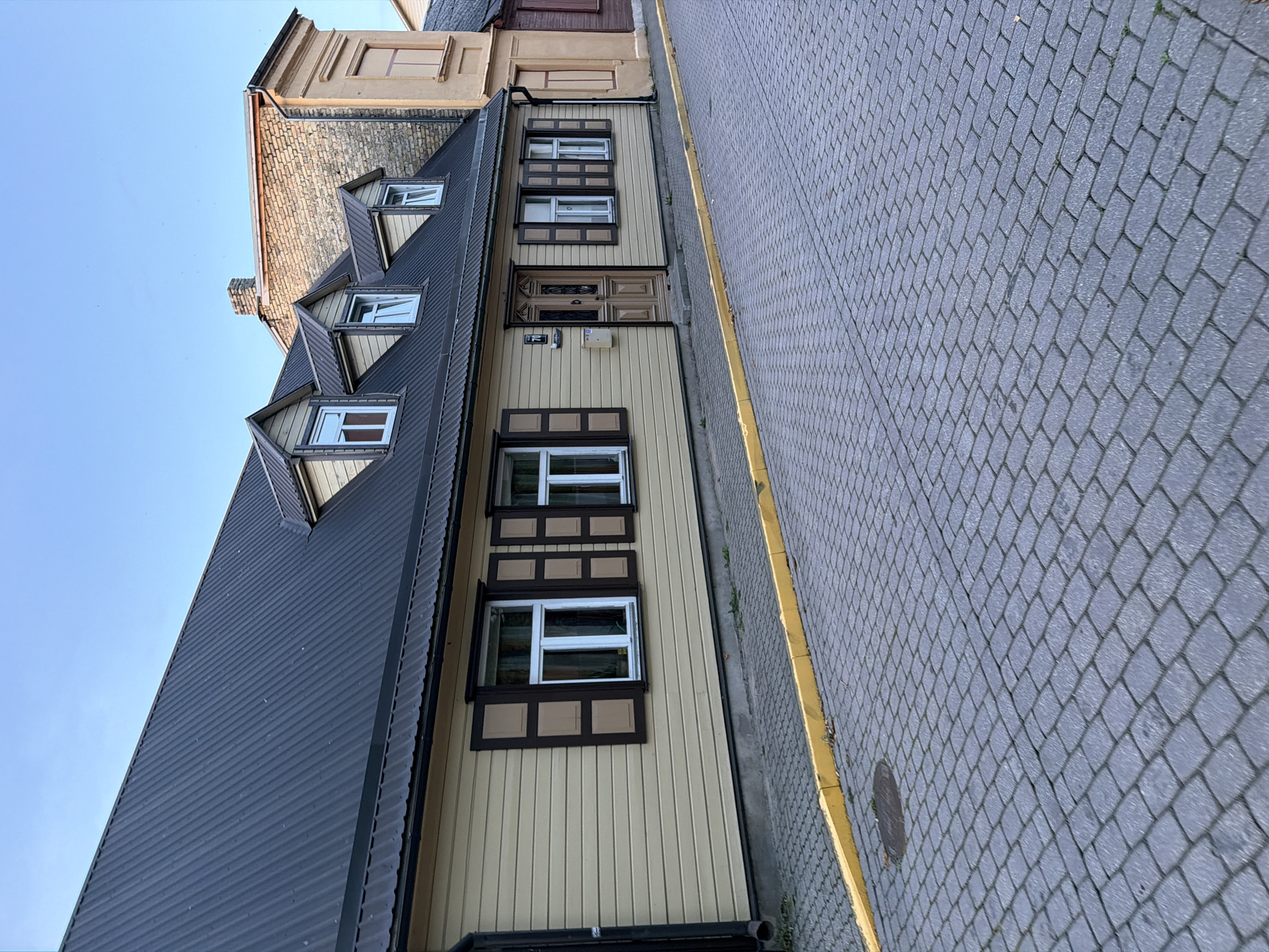 This image depicts a residential street in Ventspils, Latvia, likely from the perspective of a person standing on the sidewalk. The foreground is dominated by a cobblestone street with a distinctive yellow curb. The street is clean and appears to be in good condition.
To the right of the street is a light-colored, wooden house with a dark, corrugated metal roof. The house features multiple dormer windows on its mansard roof, each with a dark frame and white panes. The facade of the house is horizontally paneled and painted a pale yellow. Dark brown shutters with lighter brown inserts adorn the main floor windows. A wooden door, also dark brown, is visible in the center of the house, with a house number "70" above it. A mailbox is mounted to the left of the door.
Adjacent to the wooden house on the right is a taller, brick building with a lighter yellow facade on its upper section. This building has a more traditional architectural style. Further to the right, another building with a dark red facade is partially visible.
The sky above is a clear, pale blue, indicating it is likely early morning or late evening. There are no people or vehicles visible in the image. The overall impression is one of a quiet, well-maintained street in a residential area.