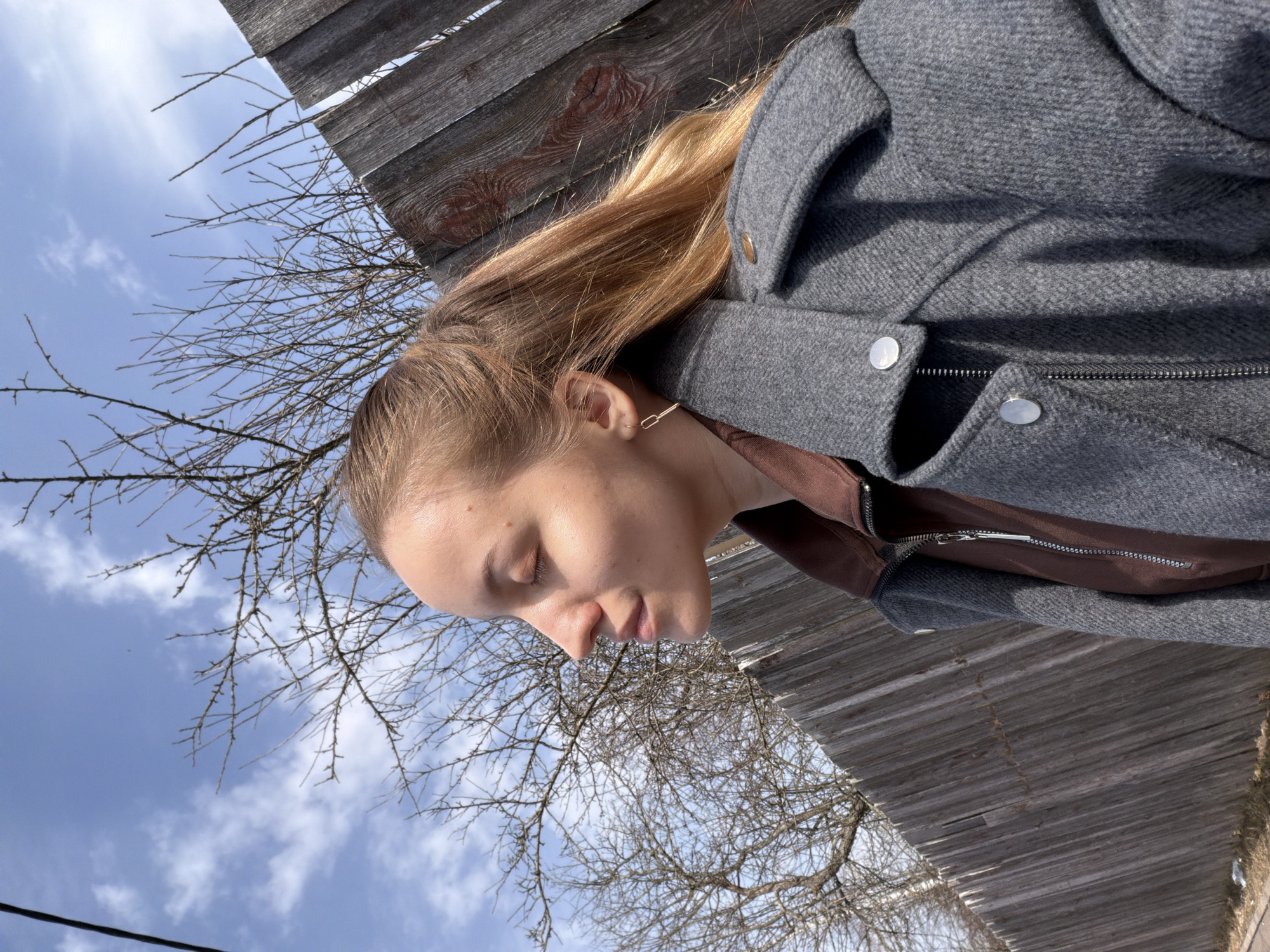 The image depicts a woman wearing a gray coat over a brown shirt, standing outdoors. The background features a wooden fence, bare tree branches, and a blue sky with scattered white clouds. The photograph was not necessarily taken in Reshetnikovo, Russia, the location is unknown.