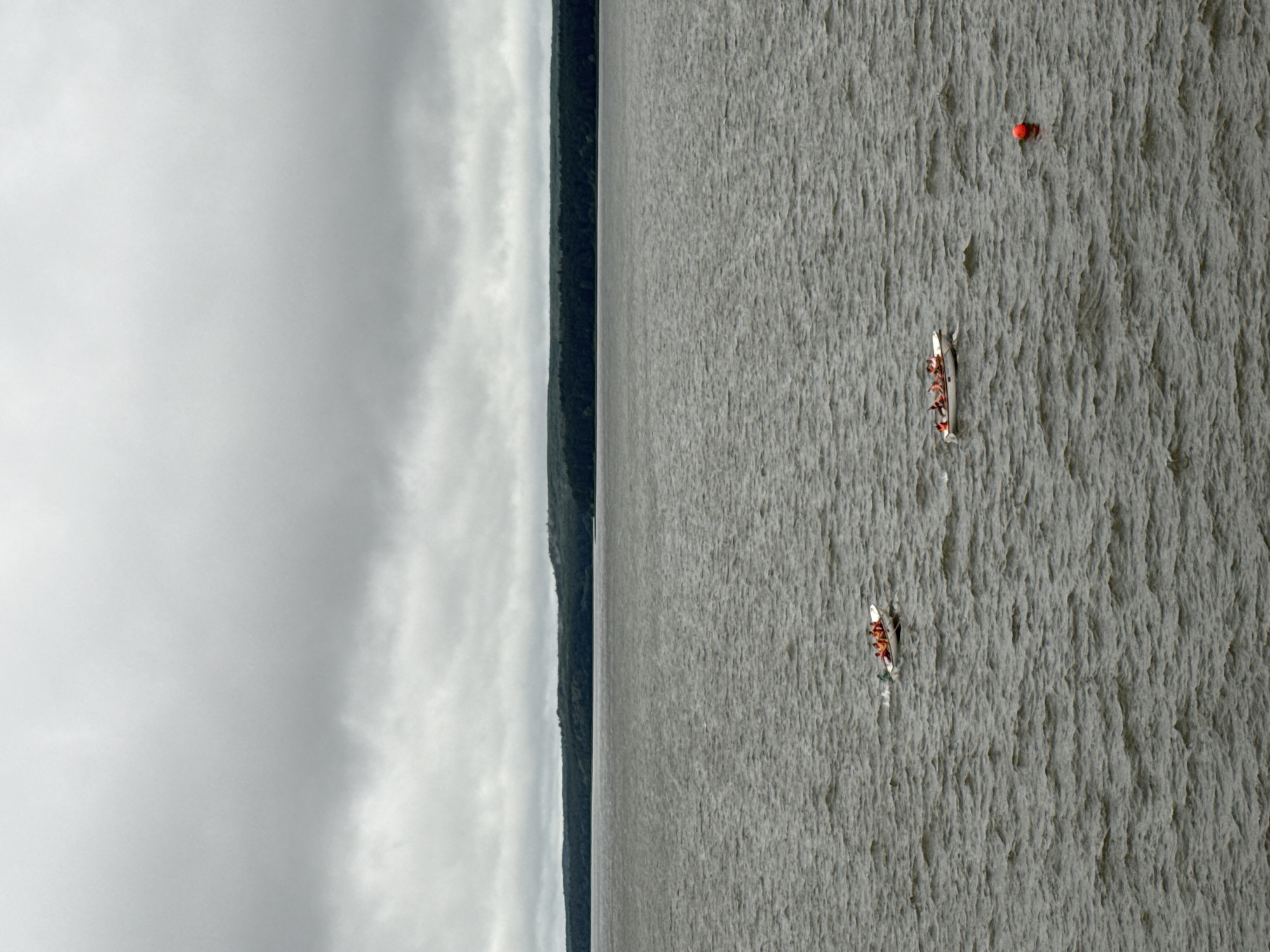 This media file shows a wide shot of a body of water, likely a lake or river, with a dark, tree-covered shoreline on the left and a cloudy sky above. The water has a textured appearance, suggesting choppy conditions. In the water, there are two canoes with people in them. The first canoe, closer to the foreground, has several individuals paddling. The second canoe is further away and also appears to be occupied by paddlers. A small red buoy is visible floating on the water in the upper right quadrant of the image. The lighting suggests it is daytime, possibly overcast given the sky. The scene depicts people engaged in a water activity, likely canoeing or kayaking, in a natural setting. There is no visible text in the image.
