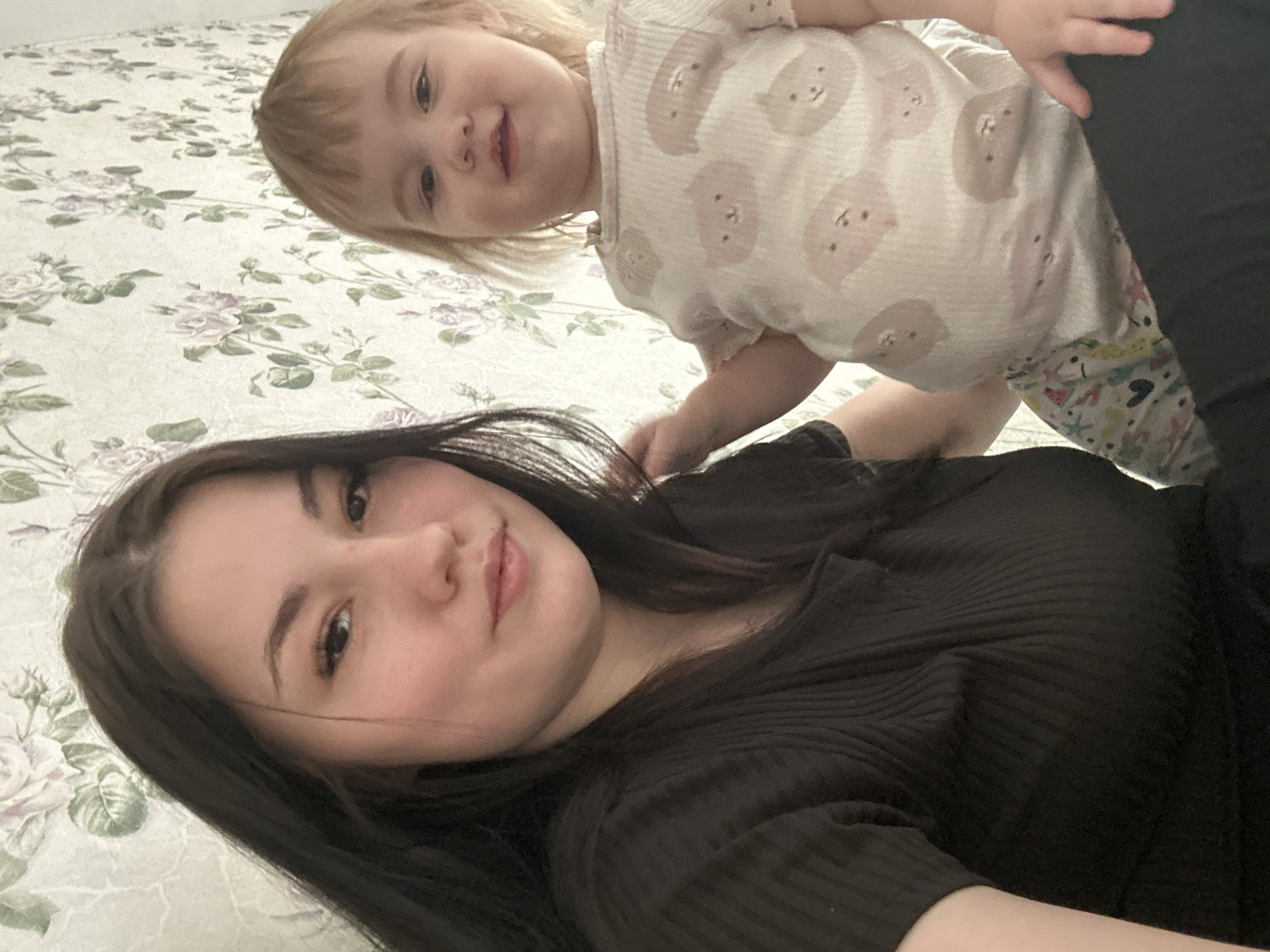 This image captures two individuals, an adult female and a young child, indoors in the city of Chaikovsky, Russia. The adult female, positioned on the left, has long dark hair and wears a dark, ribbed short-sleeved top. She looks directly at the camera with a subtle smile. The child, situated to her right and slightly lower, has light blonde hair and wears a light-colored short-sleeved top patterned with light pink and brown cartoon animal faces, and patterned pants featuring hearts and stars. The child also looks towards the camera, smiling, with its left hand resting on the adult female's upper left arm. The background consists of a light-colored wall covered in wallpaper displaying a repeating pattern of light purple rose blossoms and green foliage. The scene is illuminated by ambient, even lighting.