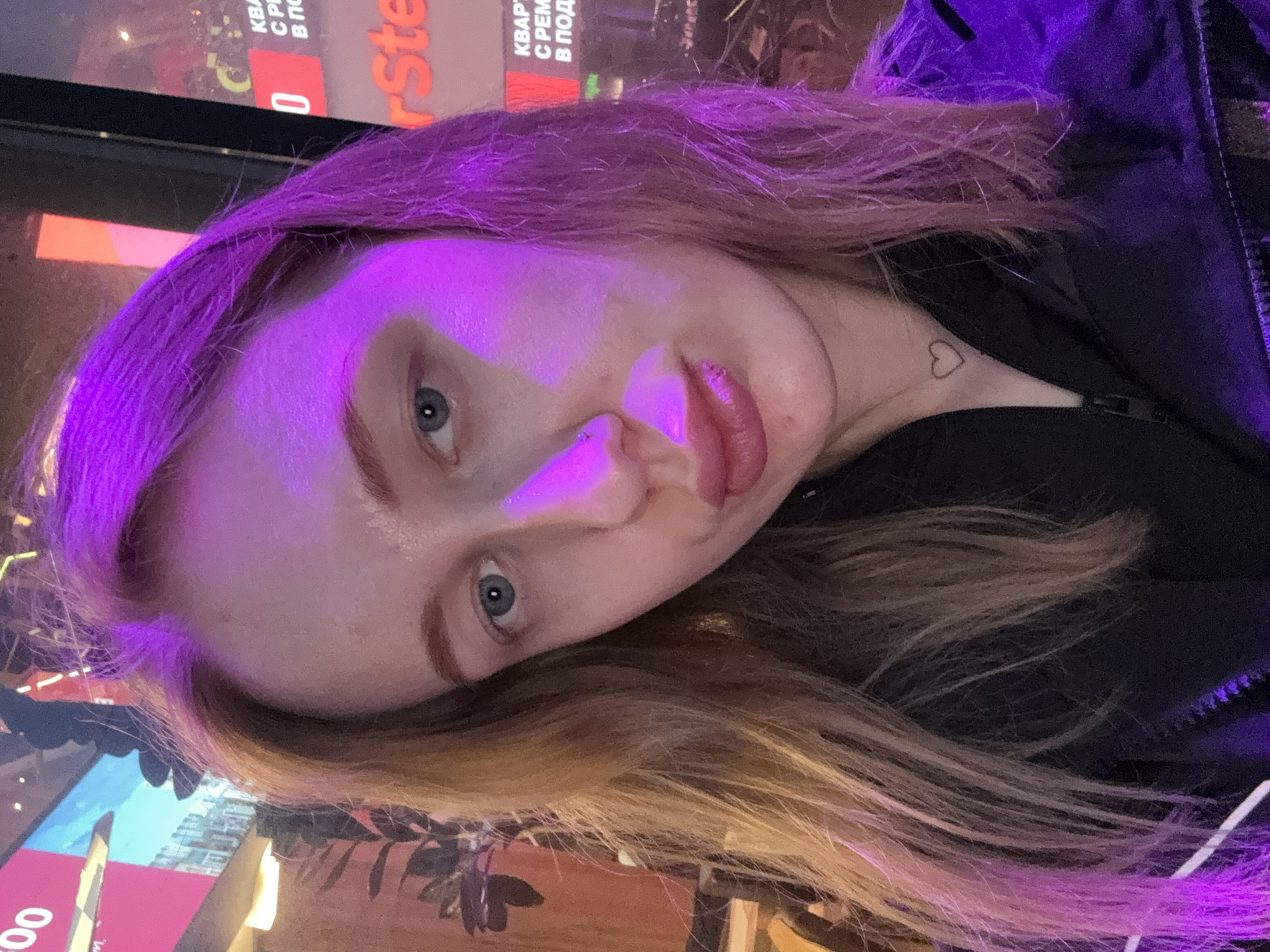 A woman with light skin and blue eyes is depicted in a close-up shot. The image is rotated counter-clockwise, casting a purple light over the upper portion of her face and hair. The woman has reddish-blonde hair that fades into lighter shades towards the ends. A small heart tattoo is visible on her neck. She is wearing a black jacket. In the background, blurred text in Russian can be seen, including the name "Ste" from a possible brand or logo. The location is likely Ol'ginskaya, Russia, based on the Russian text in the background.
