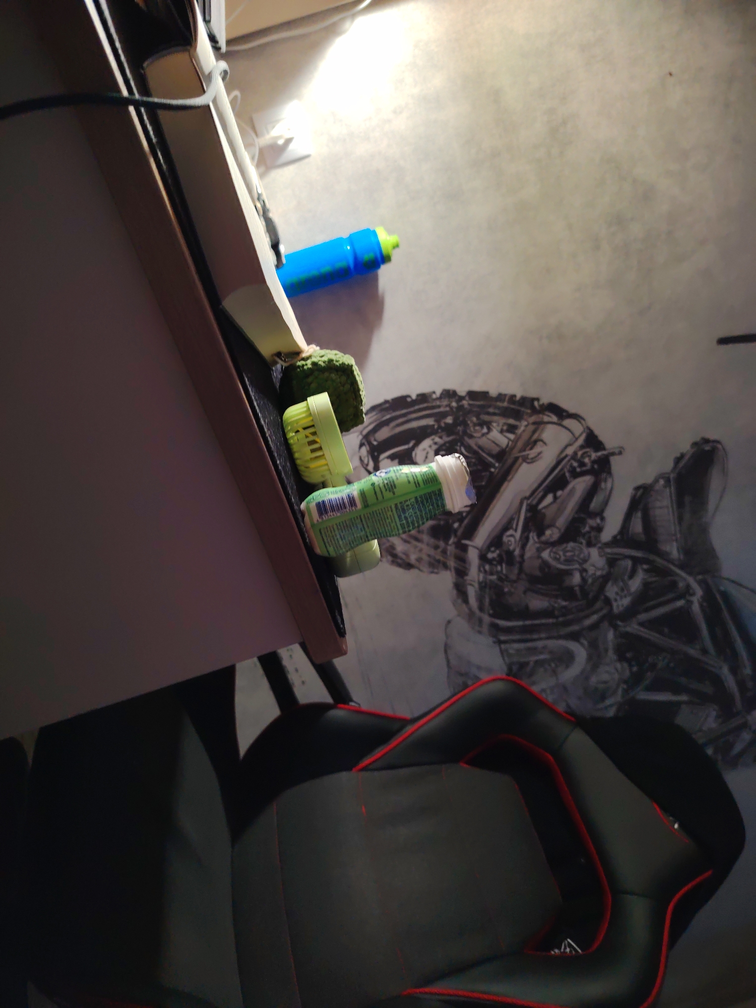 The image depicts a close-up, angled view of objects on and near a desk, with a portion of a black and red gaming chair visible in the foreground. On the desk, a bright blue water bottle with a green cap is lying down. Next to it, a green, textured object, possibly a sponge or stress ball, is visible, with a light green cylindrical object, possibly a travel-sized container of toothpaste or medication, lying on its side next to that. The background shows a gray floor with a large, detailed black and white graphic of mechanical components. The scene appears to be indoors, likely a home office or gaming setup. There are no people visible in the image, nor any indication of the specific location of "Крутые Выселки, Russia" beyond what is provided in the prompt.
