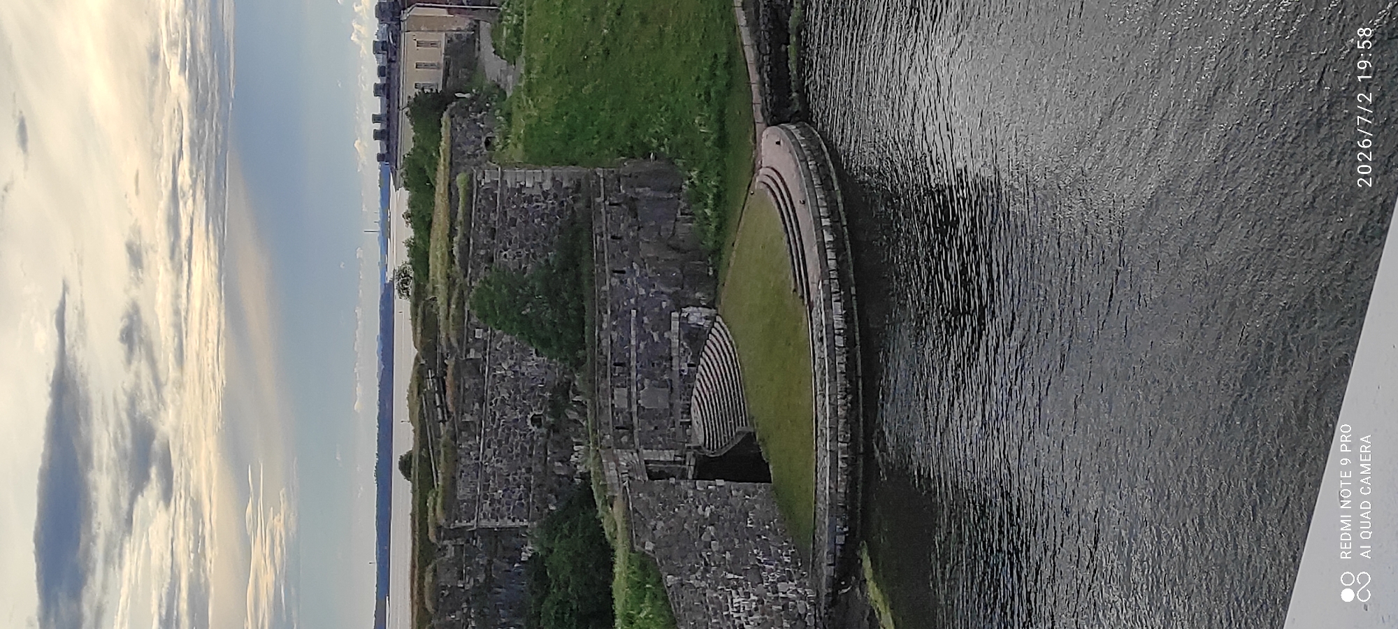 This media file depicts a historical fortress with stone walls and grassy slopes overlooking a body of water. The fortress walls are made of rough-hewn stones, suggesting an ancient or well-preserved structure. Green grass covers parts of the fortress and slopes down towards a curved, stepped embankment that meets the water's edge. The water is dark and rippled, reflecting the overcast sky.
In the background, beyond the fortress and water, are distant buildings and structures, possibly indicating a city or inhabited area. The sky is filled with a mix of bright white and grey clouds, suggesting a bright but possibly cloudy day, potentially late afternoon or early evening given the soft light.
The composition is predominantly landscape-focused, with the fortress and water as the main subjects. There are no people visible in the image. The overall impression is one of historical grandeur and natural beauty, with a sense of scale provided by the expansive sky and water.
Visible text includes a watermark at the bottom right corner: "REDMI NOTE 9 PRO AI QUAD CAMERA" and a date/time stamp: "2026/7/2 19:58". The text "2026/7/2 19:58" appears to be a future date and time. The location context provided is Helsinki, Finland, which suggests this may be a view of Suomenlinna, a sea fortress and UNESCO World Heritage site in Helsinki.