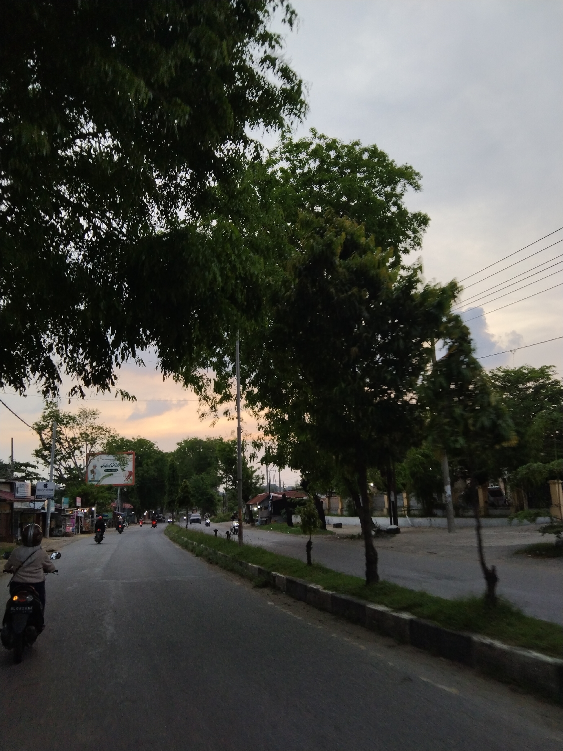 The image captures a street scene in Lhokseumawe, Indonesia, during what appears to be twilight, either early morning or late afternoon/evening. The sky displays soft hues of orange and yellow near the horizon on the left, transitioning to grey and white clouds above and to the right.
The primary subject is a paved two-lane road, stretching into the distance and populated by several vehicles. In the foreground, a person wearing a helmet and a light-colored jacket is seen from behind, riding a black motorcycle. Further down the road, other motorcycles and a white car are visible, indicating routine traffic.
Both sides of the road are lined with abundant green trees, some with dense foliage shading the scene from above, particularly on the left. On the right, a grassy verge with a concrete curb separates the road from additional trees and what appears to be an undeveloped or property area. Overhead power lines crisscross the sky on the right.
On the left side of the road, several buildings, likely shops or residences, are visible, interspersed with trees. A large billboard stands prominently above these structures. Visible text on the billboard includes "Idul Fitri" and likely other related greetings, suggesting a festive or commemorative message. Below the main text, a smaller sign reads "PALESTINE". The overall scene depicts a typical busy street with a blend of natural elements and human infrastructure.