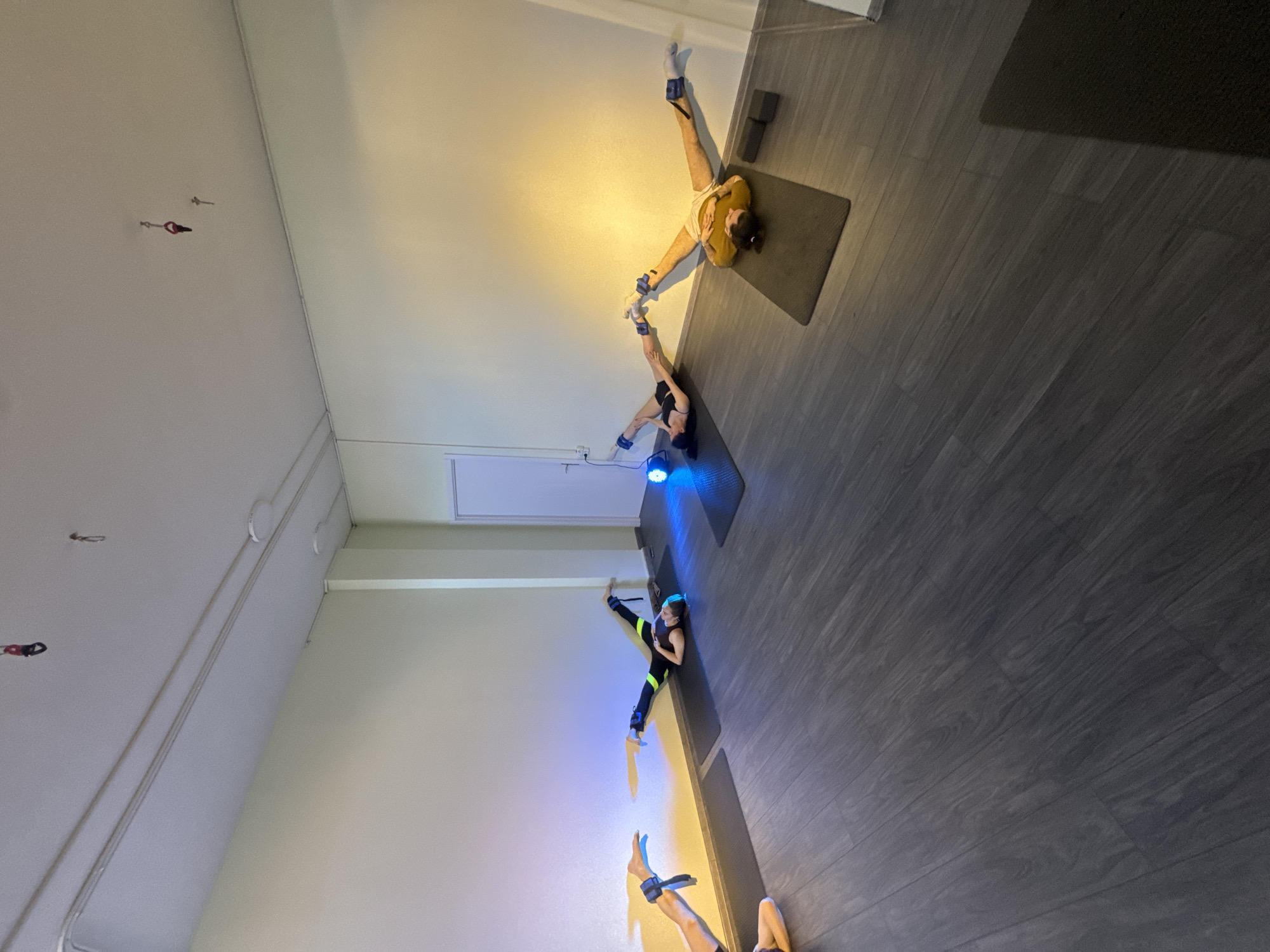 The image shows the interior of a studio, possibly for fitness or yoga, with three individuals positioned on yoga mats. Each person is wearing ankle weights and appears to be engaged in some sort of exercise or stretching routine. The room has a grey wood-like floor and white walls. The ceiling is also white and has a few small fixtures. A door is visible along one wall. The city of Unknown, Unknown, is not mentioned.