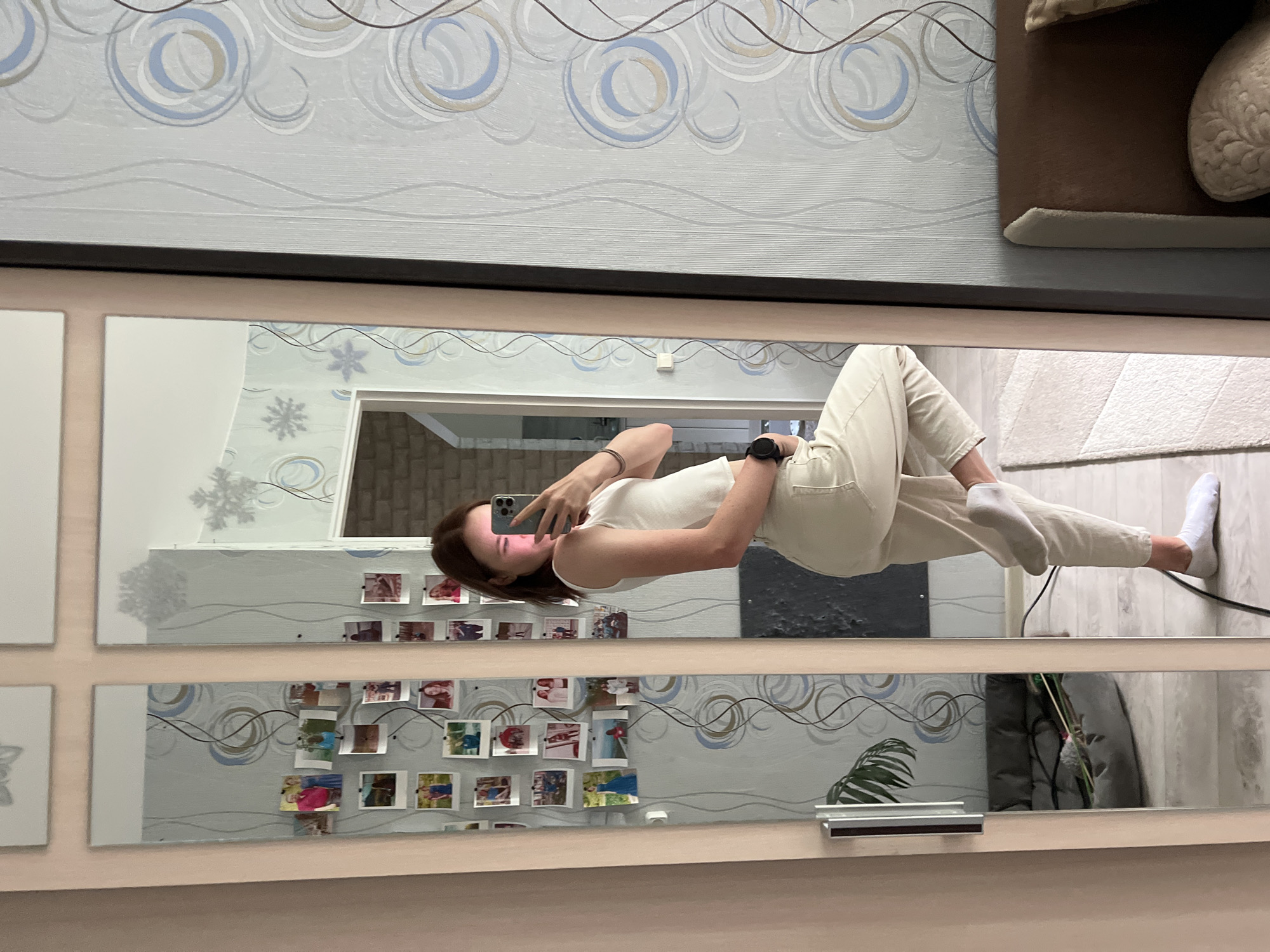 A young woman stands in front of a mirror taking a selfie with her smartphone. She is wearing a white tank top and light-colored pants. The location appears to be an indoor setting, possibly an apartment. The background includes a wall with patterned wallpaper and a mirror adorned with a collage of photographs. The reflection in the mirror reveals details of the room, including a doorway, a rug, and a potted plant. There is no specific information to determine the exact location in Chaikovsky, Russia.