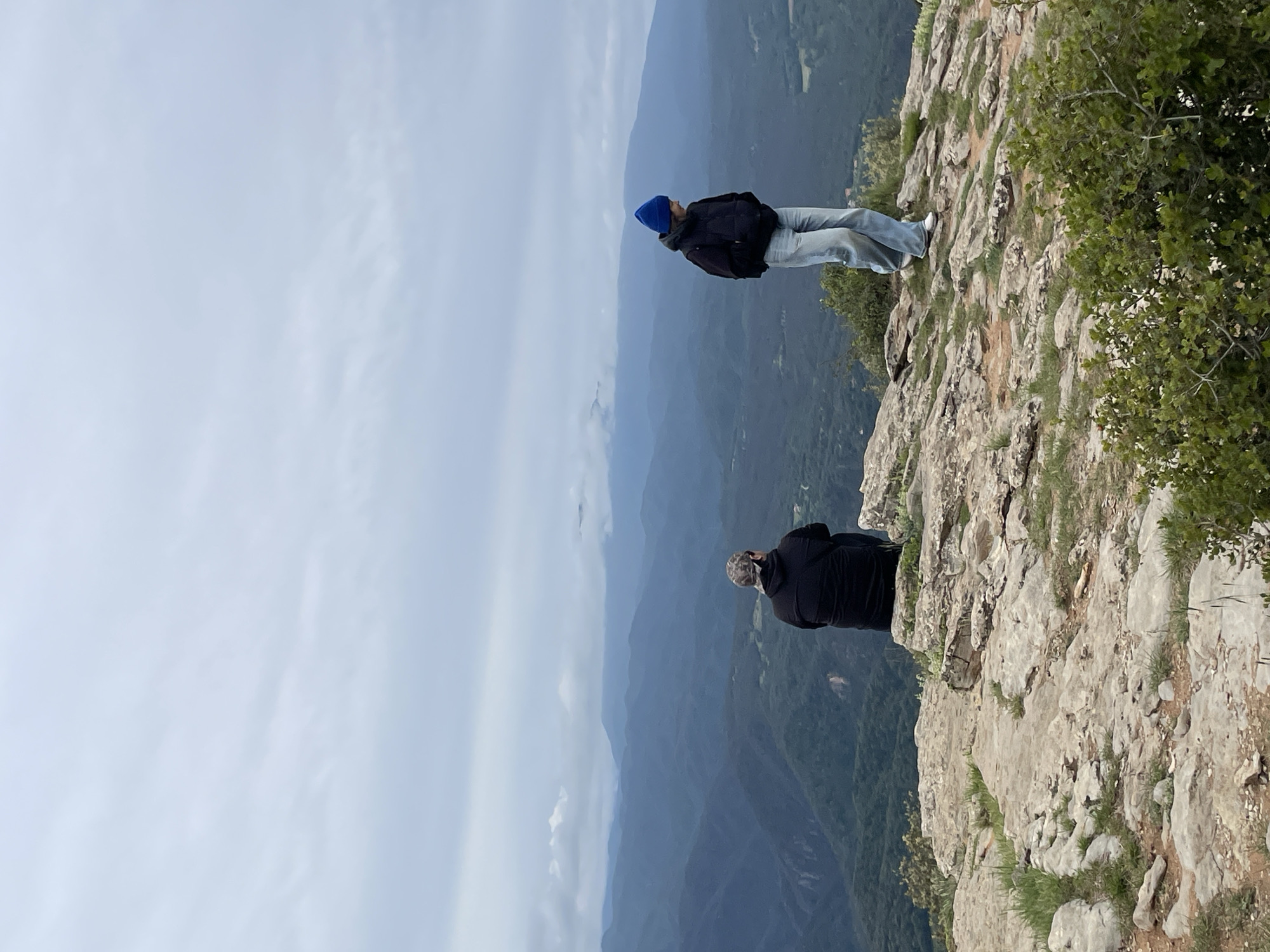 Two individuals stand on a rocky precipice, likely a cliff or mountain edge, overlooking a deep valley filled with lush green vegetation and distant mountainous terrain under a partly cloudy sky. The person closest to the camera is seated, dressed in dark clothing and a camouflage-patterned hat. The other person, farther away, is standing, wearing a blue beanie, a dark jacket, and light-colored jeans. The photograph is taken from a slightly low angle, emphasizing the height of the precipice and depth of the valley. The location is Tavertet, Spain, based on visual markers of the landscape.