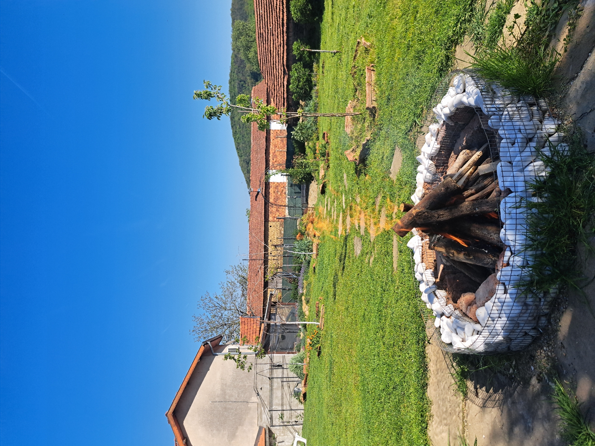 The image depicts a fire pit burning in a grassy yard. The fire pit is constructed of a wire cage filled with white rocks. Inside, logs are burning with visible flames. The yard is green, and in the background, there are several buildings with red tile roofs. In the far background, there is a tree-covered hill. The sky is a clear blue. The location is Lisets, Bulgaria.