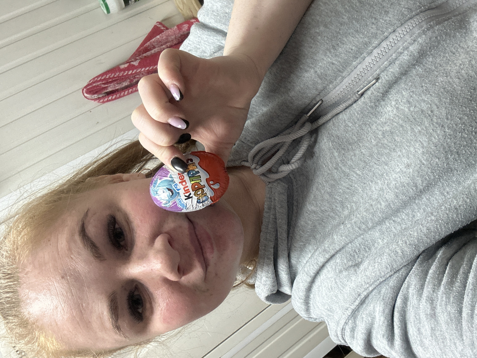 A casual, close-up indoor shot features a young woman, likely in Chaikovsky, Russia, holding a Kinder Surprise egg. She has blonde hair and is wearing a grey hooded sweatshirt with a visible zipper and drawstrings. Her right hand, adorned with manicured nails (some black, some light pink/white with a small heart design), holds the red-foiled Kinder egg, which clearly shows the "Kinder" logo and a blue/purple character. Her face, turned slightly towards the camera, reveals a faint smile or smirk, with her eyes looking forward. The background consists of white horizontal paneled walls. A folded red and white patterned cloth and a small white bottle with a green cap are visible on a ledge in the upper left corner. The image conveys a relaxed and casual atmosphere.