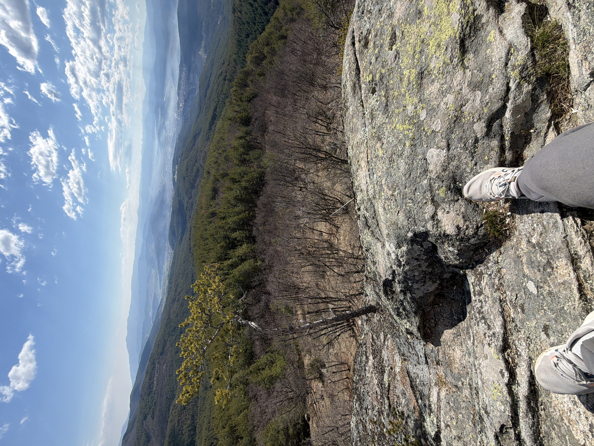 The image shows a view from a rocky precipice, likely a mountain peak, with a pair of legs and feet clad in grey athletic shoes and pants visible in the foreground. The viewpoint overlooks a forested area and a hazy valley, possibly containing a city or town. The sky above is blue with scattered white clouds. The rock surface near the viewer is textured and interspersed with patches of moss and vegetation. A solitary, stunted tree is clinging to the rock face below. Velingrad, Bulgaria is not discernible in the provided image.