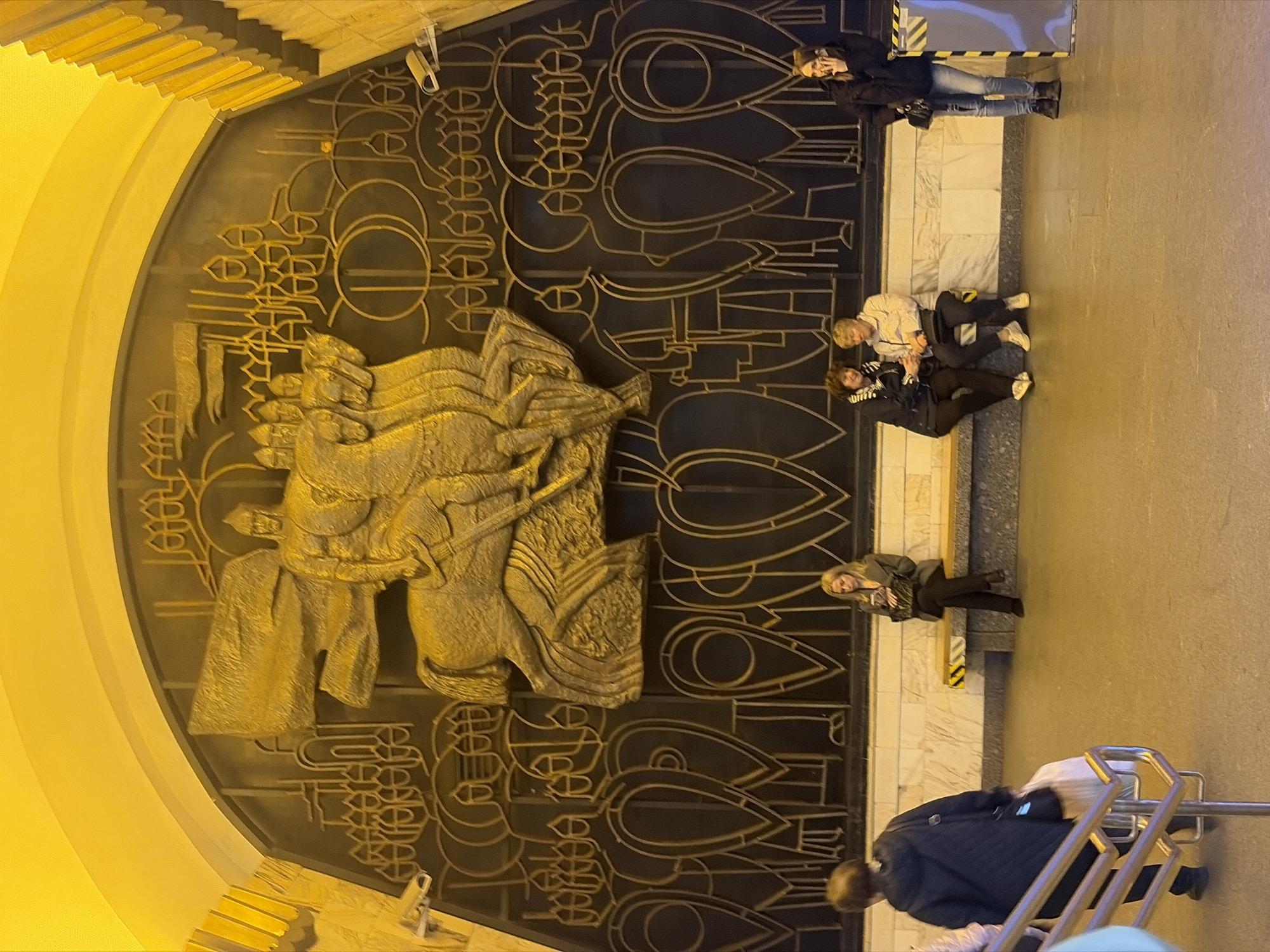 The photograph captures a scene inside the Avtovo Metro station in Saint Petersburg, Russia. The focal point is a large, semi-circular wall decoration featuring a bas-relief sculpture of a warrior on horseback, possibly representing a historical figure or allegorical symbol. The background of the artwork is adorned with stylized geometric patterns, possibly representing figures or shields.
Several individuals are present in the scene. Two people are seated on a bench to the right, while another person stands nearby. A woman in a dark coat walks towards the foreground with some kind of luggage cart. Another person is standing and leaning against a wall.
The architectural environment includes yellow arches on the left and a marble or stone wall on the right. The overall lighting is bright and typical of an underground metro station.