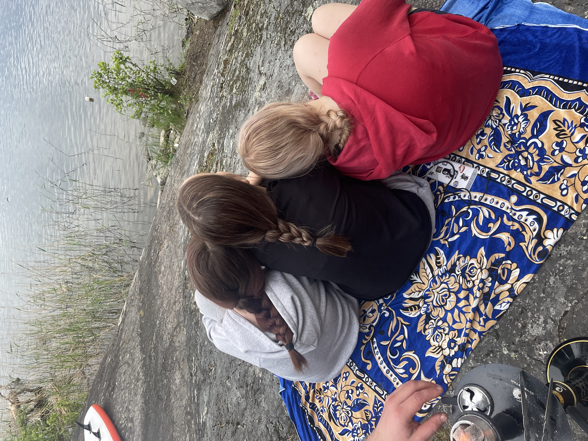 The image displays three individuals seated from behind on a patterned blue and gold blanket, positioned on a rocky shore next to a calm body of water. The scene is set outdoors, under an overcast sky. While the precise location within Yarkoe, Russia, is not discernible from the visual information, the environment suggests a natural lakeside or riverside setting.
Three young females are visible. The leftmost individual wears a light grey hooded sweatshirt and has dark brown hair styled in a single braid. She leans against the middle individual, who is wearing a black top and has medium brown hair in a single braid. The rightmost individual wears a red hooded sweatshirt and has blonde hair in a single braid; she also leans against the middle individual.
In the lower-left foreground, a partially visible hand, belonging to a fourth person, is positioned near a black hookah. The hookah's metal bowl contains what appears to be lit charcoal. To the far left on the rocky ground, a white and orange inflatable board with two black fins is partially visible.
The shoreline along the body of water is characterized by tall reeds and a small green bush. Several pieces of refuse, including plastic debris and an object resembling a plastic bottle, are scattered near the water's edge and within the water. On the patterned blanket, near the rightmost individual, lies a white mobile phone with a dark, stickered case and an attached cable. A plain blue towel is also visible beneath the rightmost individual on the blanket. The ground consists of greyish, irregular rock and soil.