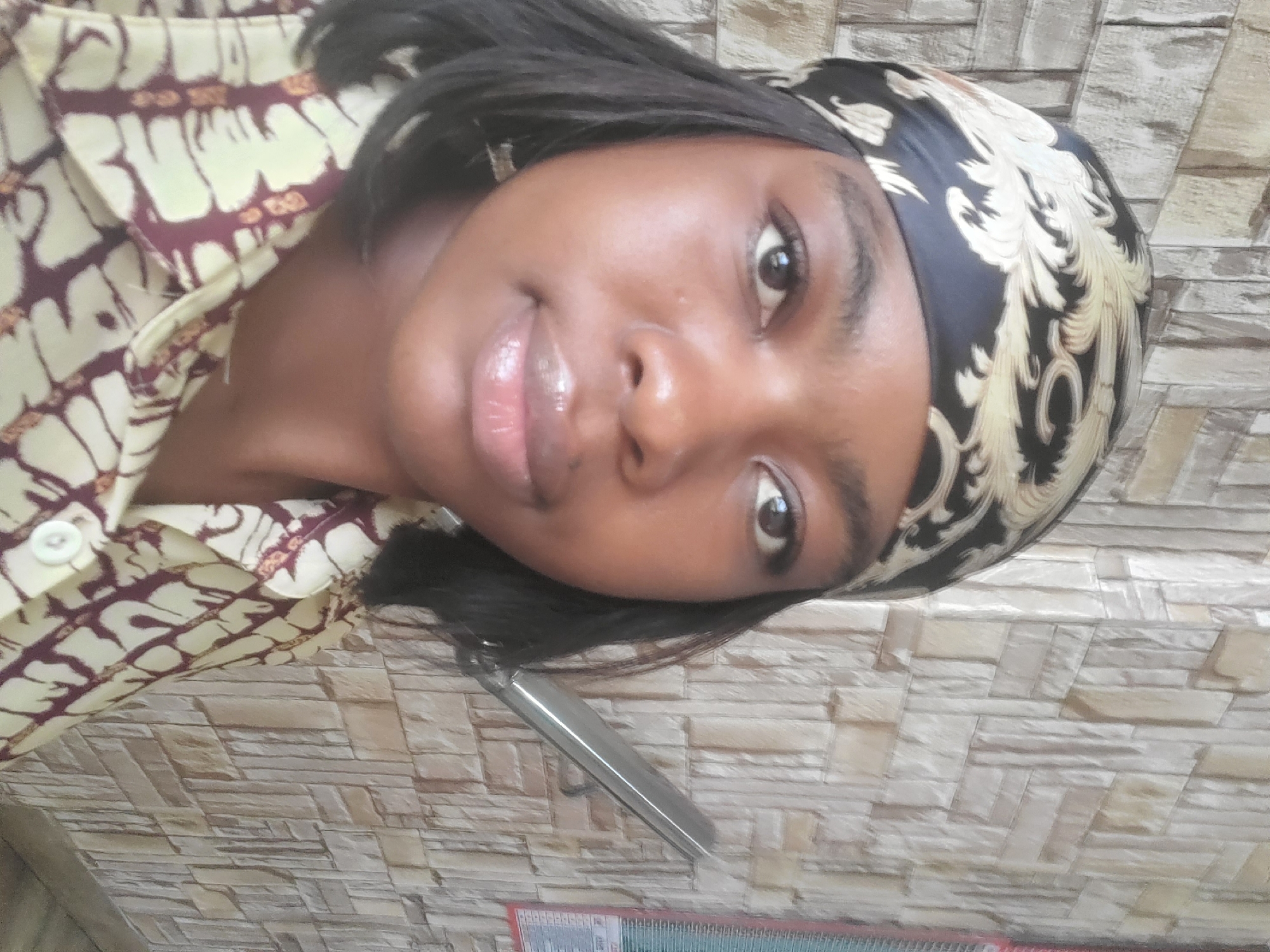 A young woman with dark skin and black hair is centered in the frame. She is wearing a patterned shirt and a black headscarf with a gold design. Her lips appear glossy. The background consists of a textured wall resembling stacked stone. A silver object is visible near her hair, and a calendar is at the bottom of the frame. The image is rotated to the side. There is no visual information to confirm the location of the image within Gwagwada, Nigeria.