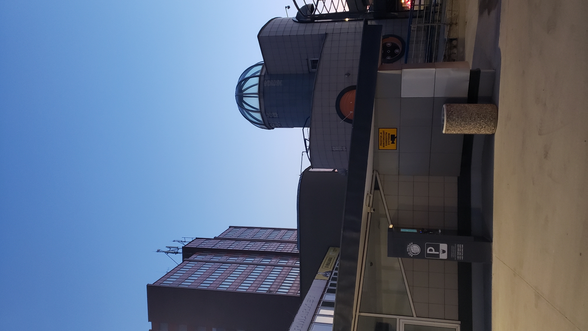 The image shows the exterior of a multi-story building with a distinctive domed glass roof structure in Prešov, Slovakia. The building features a modern architectural style with a dark gray tiled facade. A tall, dark rectangular building with rows of windows is visible to the left. In the foreground, a dark gray pillar with signage displays parking information and a "P" symbol. A yellow sign with a camera icon and text in Slovak, indicating surveillance, is mounted on the building wall. A cylindrical concrete trash receptacle stands to the right of the pillar. The sky above is a clear, pale blue, suggesting it is either early morning or late evening. The overall scene depicts an urban environment with commercial or public facilities.