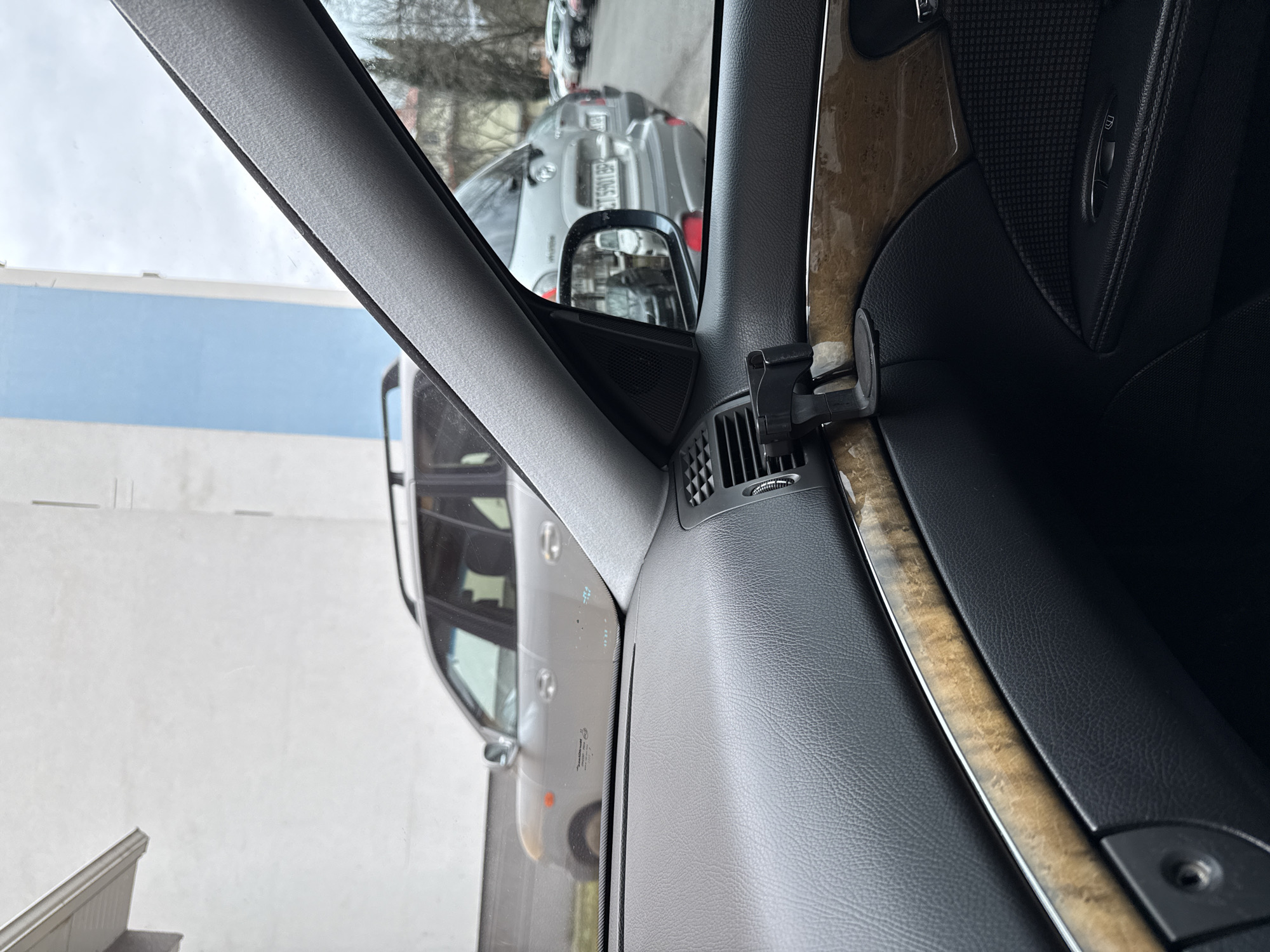 The image shows the interior of a car, specifically the driver's side door panel and a view through the front window. The door panel is a mix of gray plastic, wood trim, and black leather. An air vent is visible on the door panel near the side mirror mount, where a black car phone holder is attached. Through the front window, other cars are visible parked on the street, a gray building, and some bare trees. A license plate is visible, with the characters "5901BP". From that detail, we can infer the location is Stara Zagora, Bulgaria.