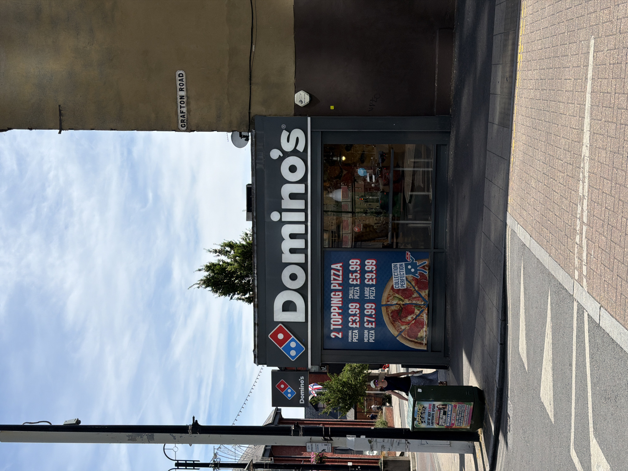 A Domino's Pizza restaurant is visible on a street corner under a partly cloudy sky. The building has a dark gray facade with the Domino's logo prominently displayed. A large window advertises a "2 Topping Pizza" deal with prices for personal, small, medium, and large sizes. A woman in a dark top and a patterned skirt is walking away from the camera towards the left side of the frame, carrying a light-colored bag. To the left of the Domino's, a green metal bin with posters on it is situated on the sidewalk. In the background, a street sign reads "CRAFT ON ROAD". The street in the foreground has a pedestrian crossing with white markings.
