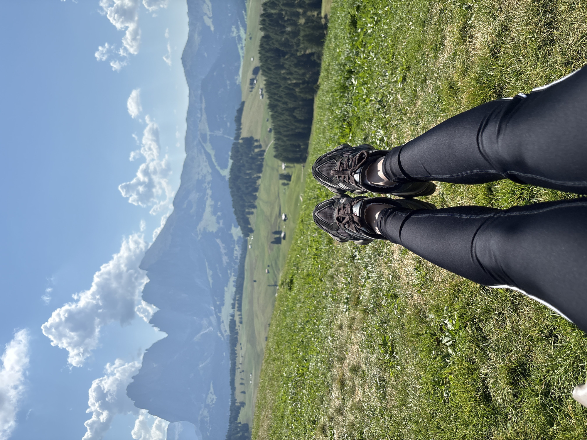 A first-person perspective shot captures a person's legs, clad in black leggings and dark athletic shoes, resting on a vibrant green grassy hillside. The background unfolds into a breathtaking panorama of the Dolomites mountain range, showcasing jagged peaks piercing a clear blue sky dotted with fluffy white clouds. Rolling green hills, dotted with sparse trees and small buildings, descend towards a dense forest in the mid-ground. The overall scene depicts a serene moment in the picturesque landscape of Kastelruth - Castelrotto, Italy.