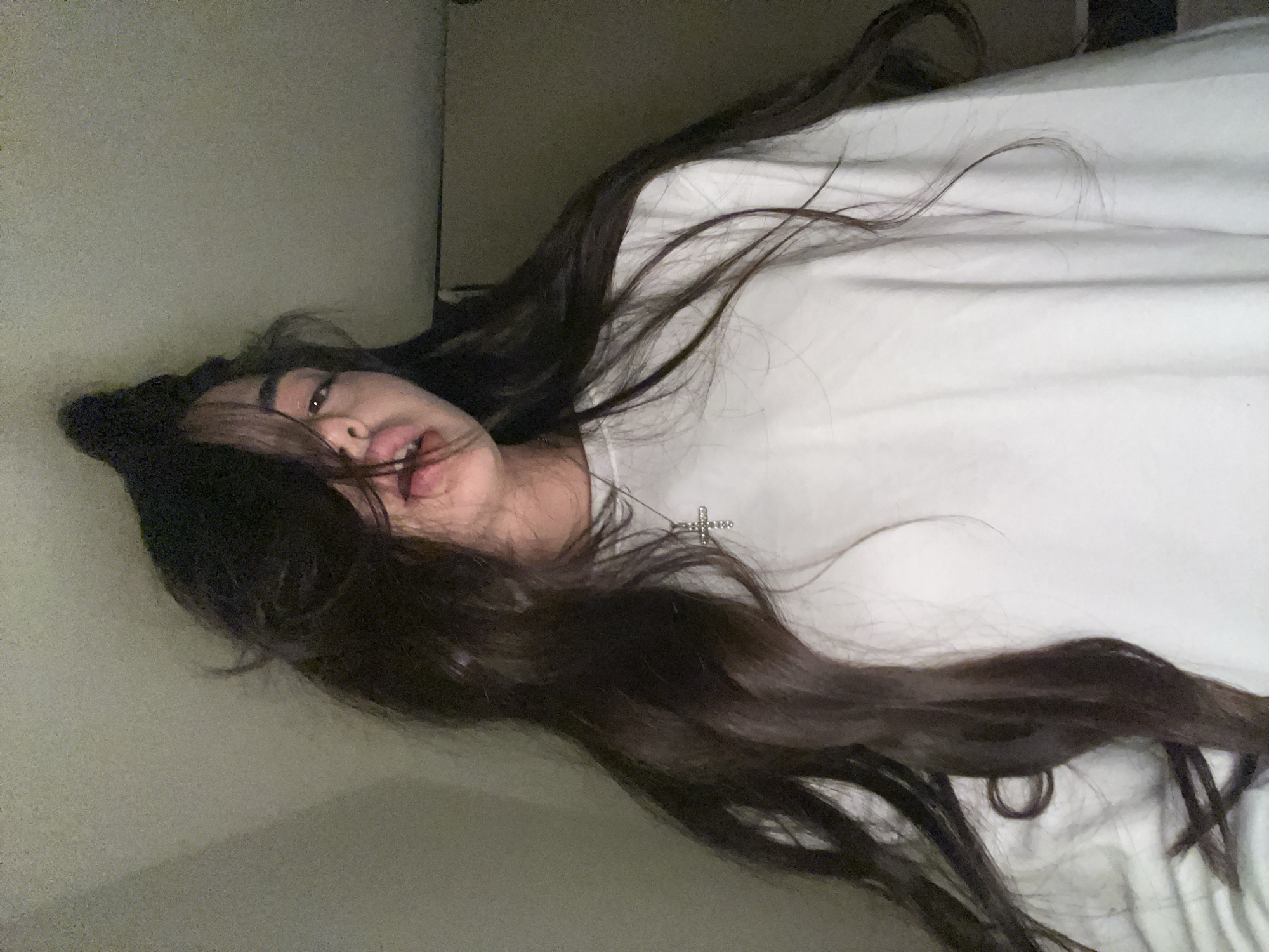 A young woman with long, dark hair is pictured from the chest up, looking at the camera. Her hair is partially covering her face, with strands falling over her eyes and forehead. Her mouth is slightly open, with her tongue visible. She is wearing a white crewneck sweatshirt and a silver cross necklace. The background appears to be a plain, light-colored wall. The image is likely a selfie taken in an indoor setting. There is no visible indication of the location in Chotěšov, Czechia.