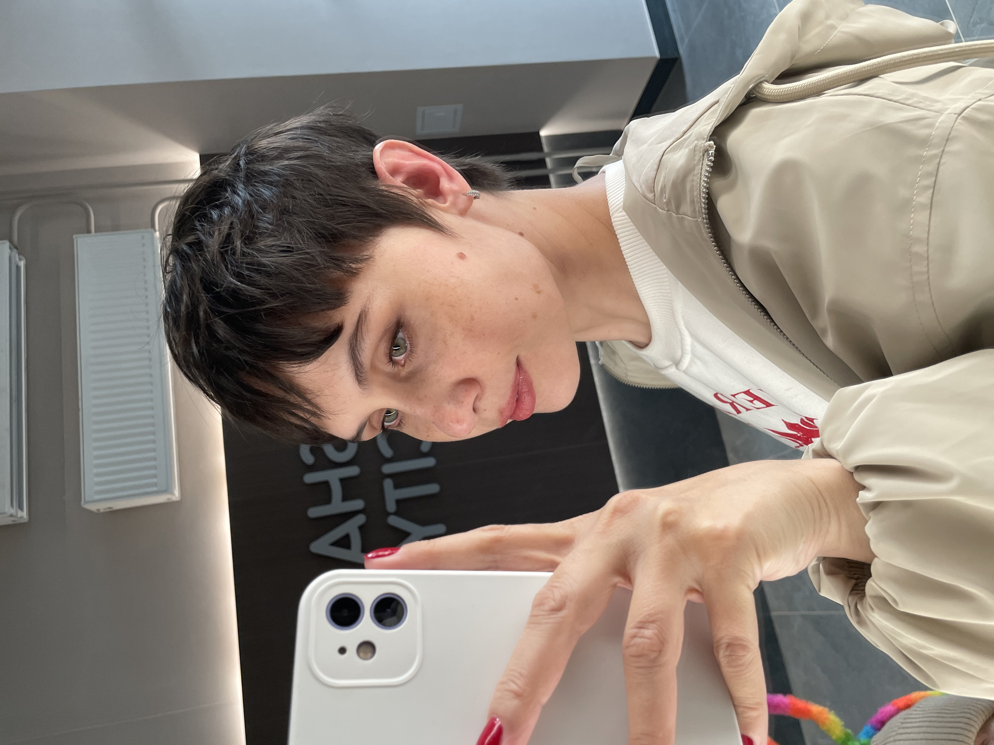 A close-up, selfie-style photograph depicts a person with short, dark hair and light eyes, holding a white smartphone with a dual-lens camera. The person is fair-skinned with freckles. They are wearing a white sweatshirt and a light beige jacket. The person's fingernails are painted red.
In the background, there's a wall with vertical lighting fixtures and black paneling with grey lettering that appears to read "CITY". The location is specified as Izhevsk, Russia.