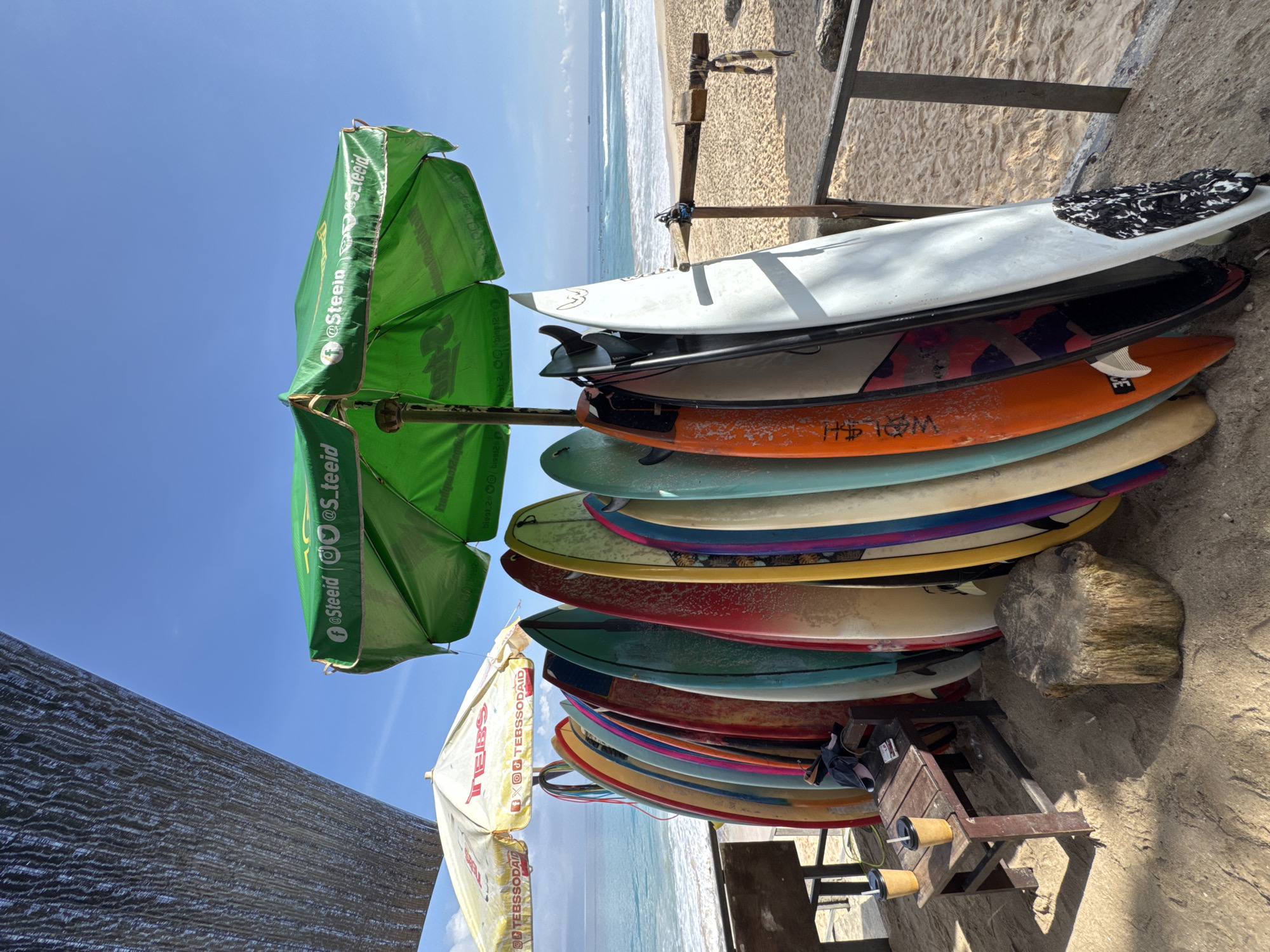 The image depicts a stack of at least 12 surfboards leaning against a wooden rack on a sandy beach. The boards display a variety of colors and designs. Two green and white umbrellas are positioned on either side of the rack. The sky is a clear blue. In the background, the ocean meets the shoreline. Based on visual clues and information available, this scene is likely located in Kuta Selatan, Indonesia, which is a known surfing destination.