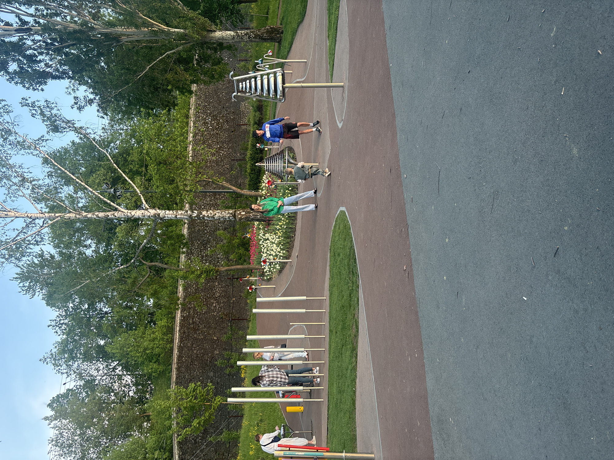 The image depicts an outdoor musical playground. Three musical instrument stations are visible, including metal xylophone-like instruments and metal chimes. Several people are interacting with the instruments, including adults and children. The playground is located along a walkway with a stone wall in the background. The location is most likely a park in the city of Perm, Russia.