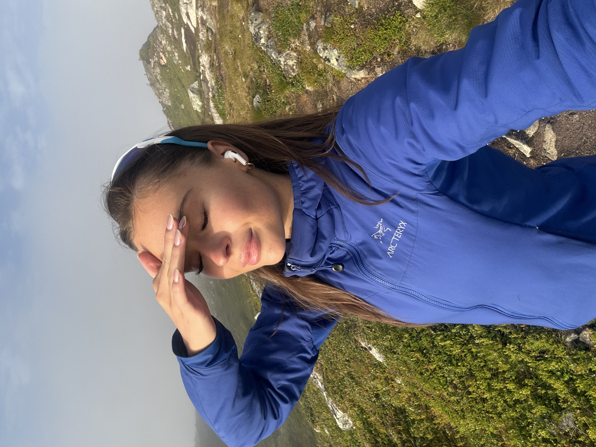 A young woman with long brown hair is posing for a selfie outdoors on a rocky, grassy hillside with mountains in the background. She is wearing a blue Arc'teryx jacket, a blue and white headband, and white wireless earbuds. Her right hand is raised to shield her eyes from the sun as she looks off to the side. The sky is overcast, with fog obscuring the distant mountains. The location appears to be a hiking trail in the mountains surrounding Harstad, Norway.