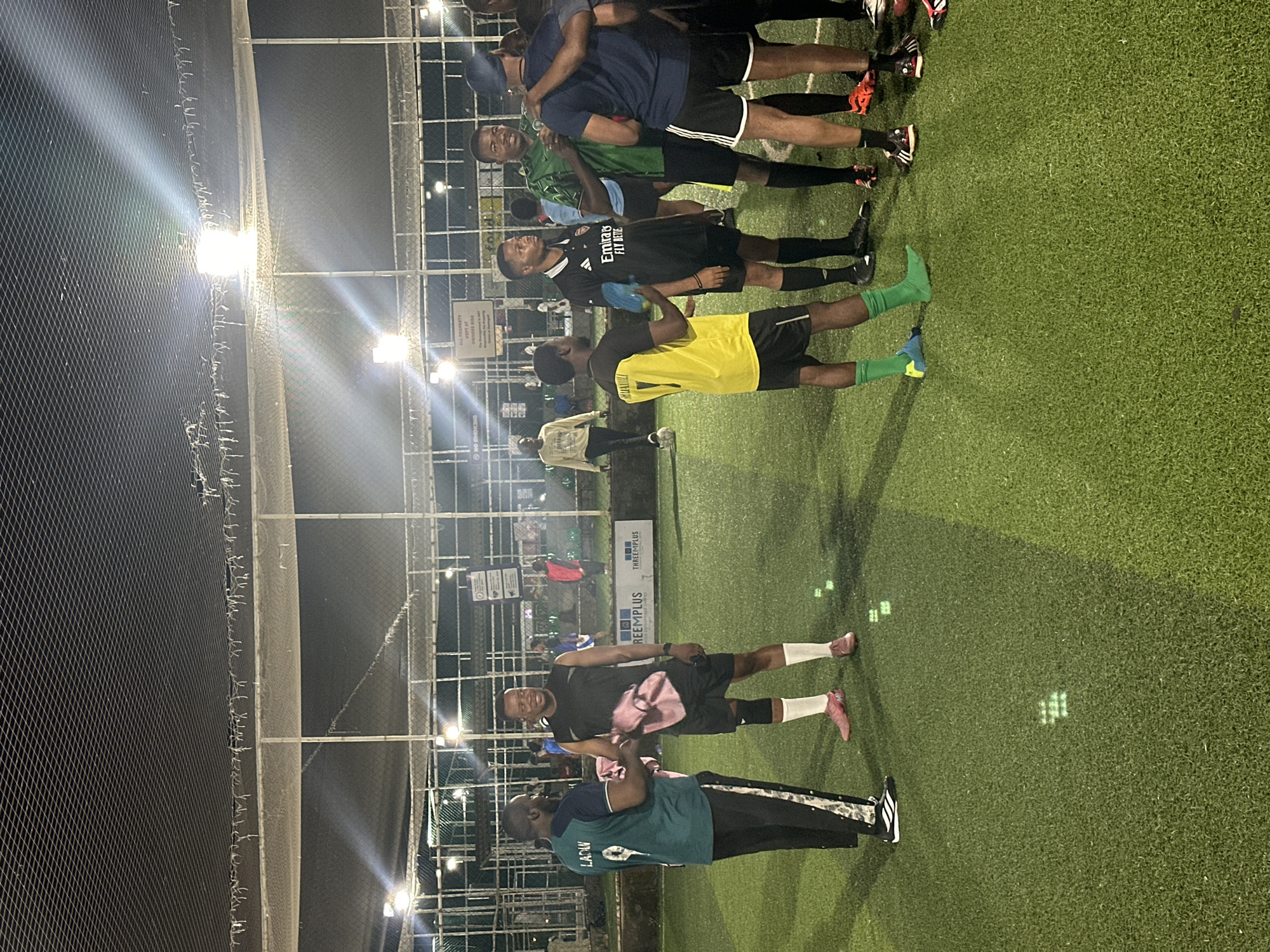 A group of young men are gathered on a green artificial turf football field, likely in Abuja, Nigeria, at night. The field is illuminated by overhead floodlights, creating bright spots and shadows. Many of the men are wearing football kits, some with jerseys and shorts, others in tracksuits. Some are wearing cleats. One man in a green and black tracksuit is shaking hands with another man in a black sleeveless top and black shorts, who is holding a pink towel. Other men are standing around, talking and observing. In the background, there are fences and netting, and glimpses of buildings and other structures, suggesting an urban or semi-urban setting. There are signs on the fence with logos and text, one of which reads "THREEPLUS". The overall atmosphere appears to be casual and social, possibly before or after a game.