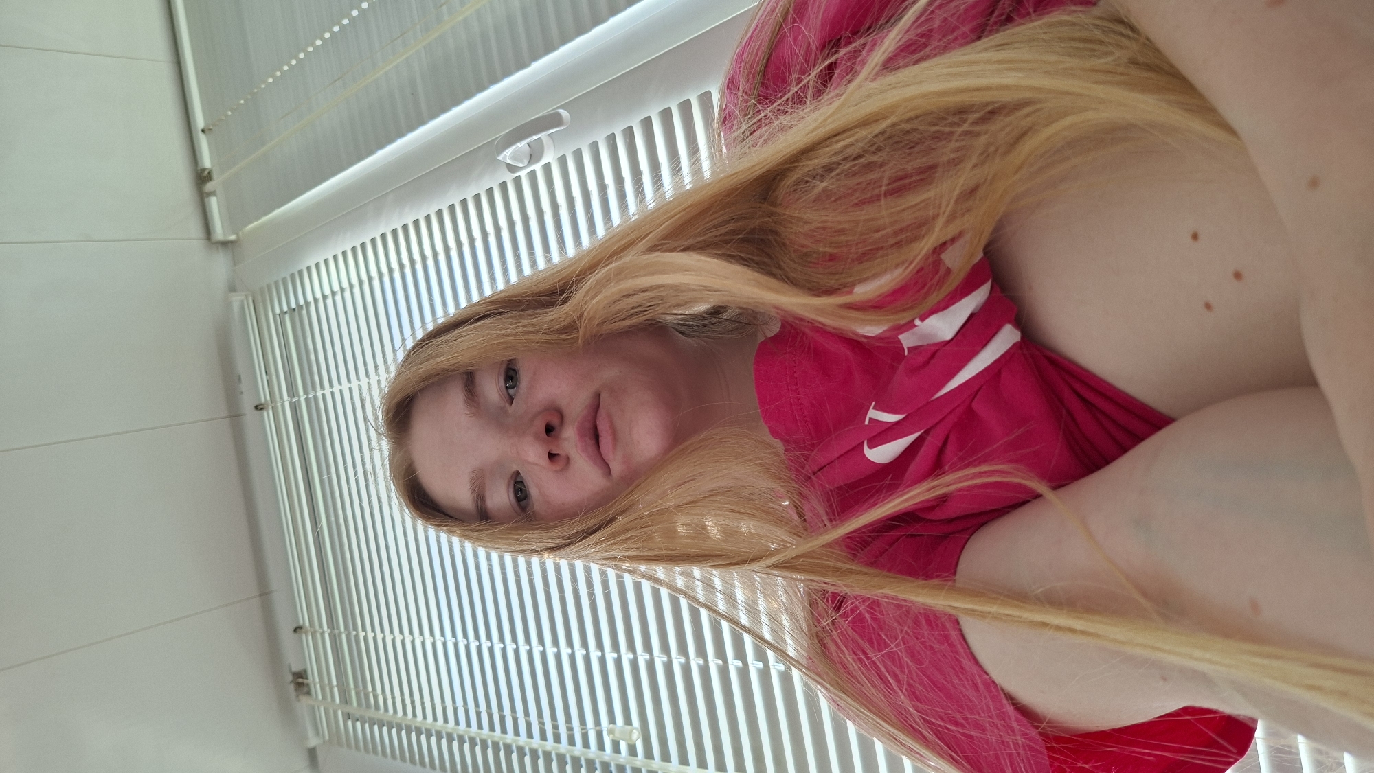 A low-angle shot captures a woman with long, blonde hair wearing a pink top with white stripes. She is positioned above the camera, gazing downward with a neutral expression. The background features white Venetian blinds. The location is in a city of Chaikovsky, Russia.