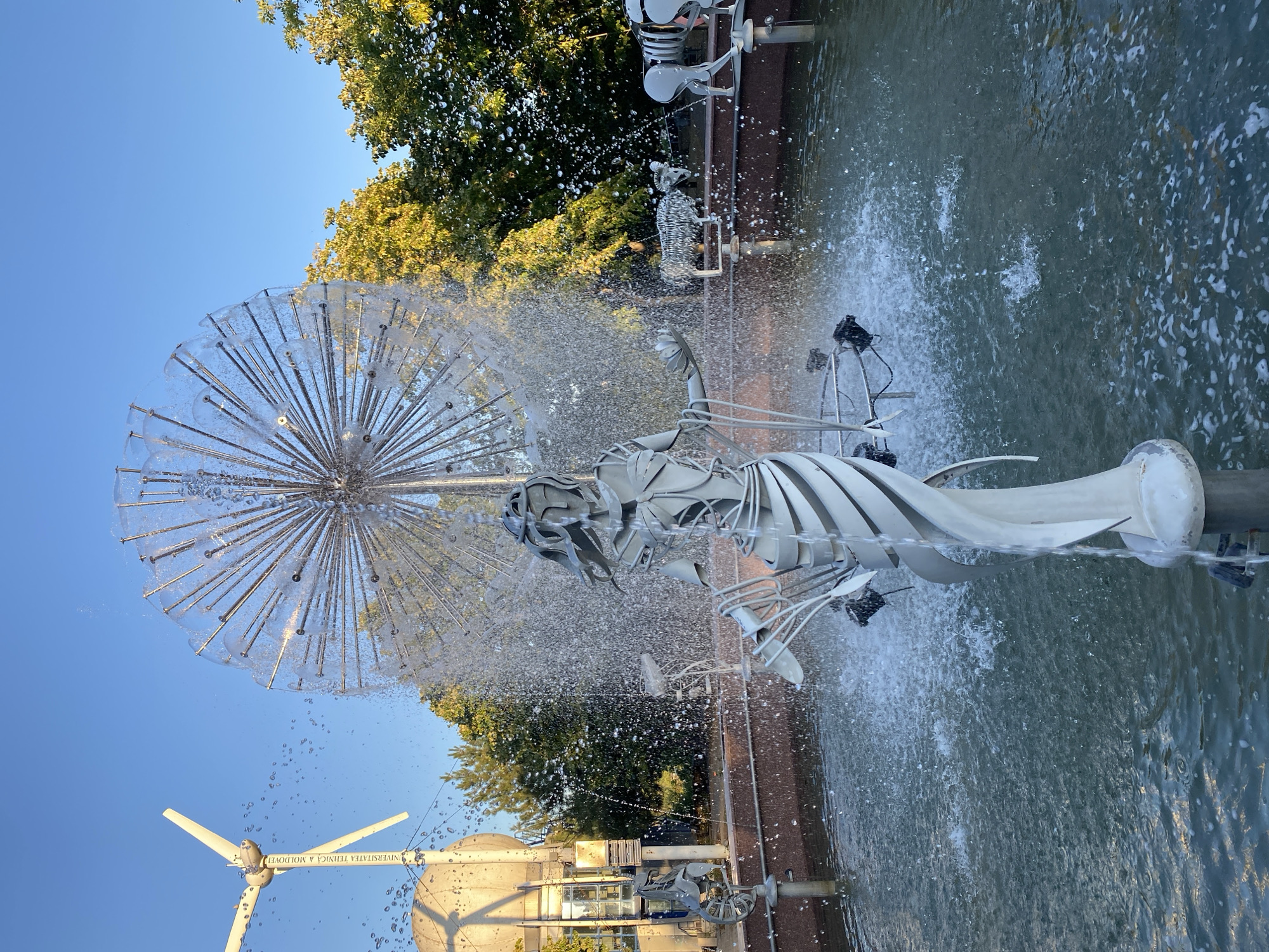 The image shows a fountain in the city of Chișinău, Moldova, specifically on the campus of the Technical University of Moldova. The fountain features a central metallic sculpture of a woman with flowing drapery and an elaborate, spiky sphere above her that sprays water. In the background, a wind turbine with visible text reading "UNIVERSITATEA TEHNICĂ A MOLDOVEI" stands against a clear blue sky. Scattered around the fountain are several metallic animal sculptures, including a ram and a horse-like creature. Trees with green and yellow foliage are visible behind the fountain and sculptures, indicating an outdoor setting. The water in the fountain is actively spraying, creating a dynamic and wet environment. The lighting suggests it is late afternoon or early evening, with sunlight casting a warm glow on the scene.