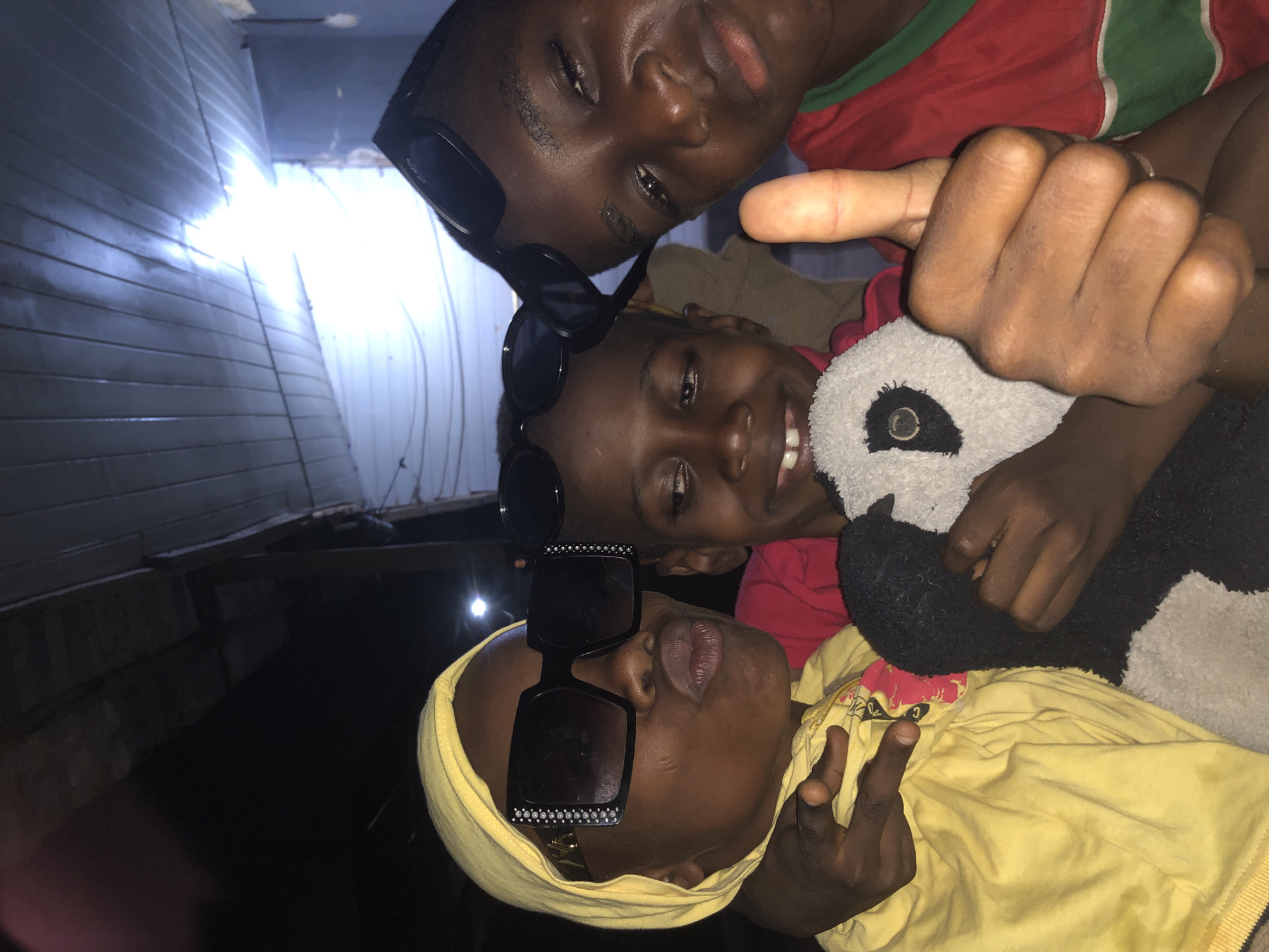 In Ado, Nigeria, three young people are captured in a close-up selfie. The person on the left wears a yellow hoodie and large, embellished sunglasses, with their lips puckered. To their right, a child in a red and green shirt, also wearing sunglasses, smiles and holds a stuffed panda. The third person, on the far right, is partially visible, giving a thumbs-up. The background appears to be an indoor or covered outdoor space with a corrugated metal ceiling illuminated by a bright light source, suggesting a modest dwelling or makeshift shelter. The overall ambiance is casual and youthful.
