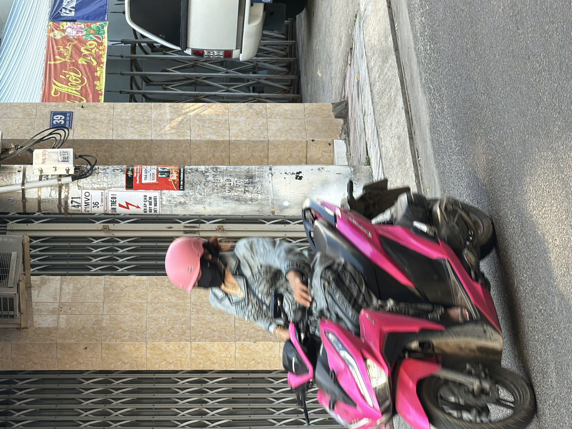 A woman wearing a pink helmet and a black mask rides a pink scooter in Nha Trang, Vietnam. She is wearing a grey patterned long-sleeved shirt and dark pants. The scooter is moving, with motion blur visible on the wheels. Behind her, a light-colored building with beige tiles and metal accordion gates is visible. A utility pole stands next to the building, with various signs attached to it, including one with the number 39. To the right of the building, a white pickup truck is partially visible. The scene appears to be on a city street.