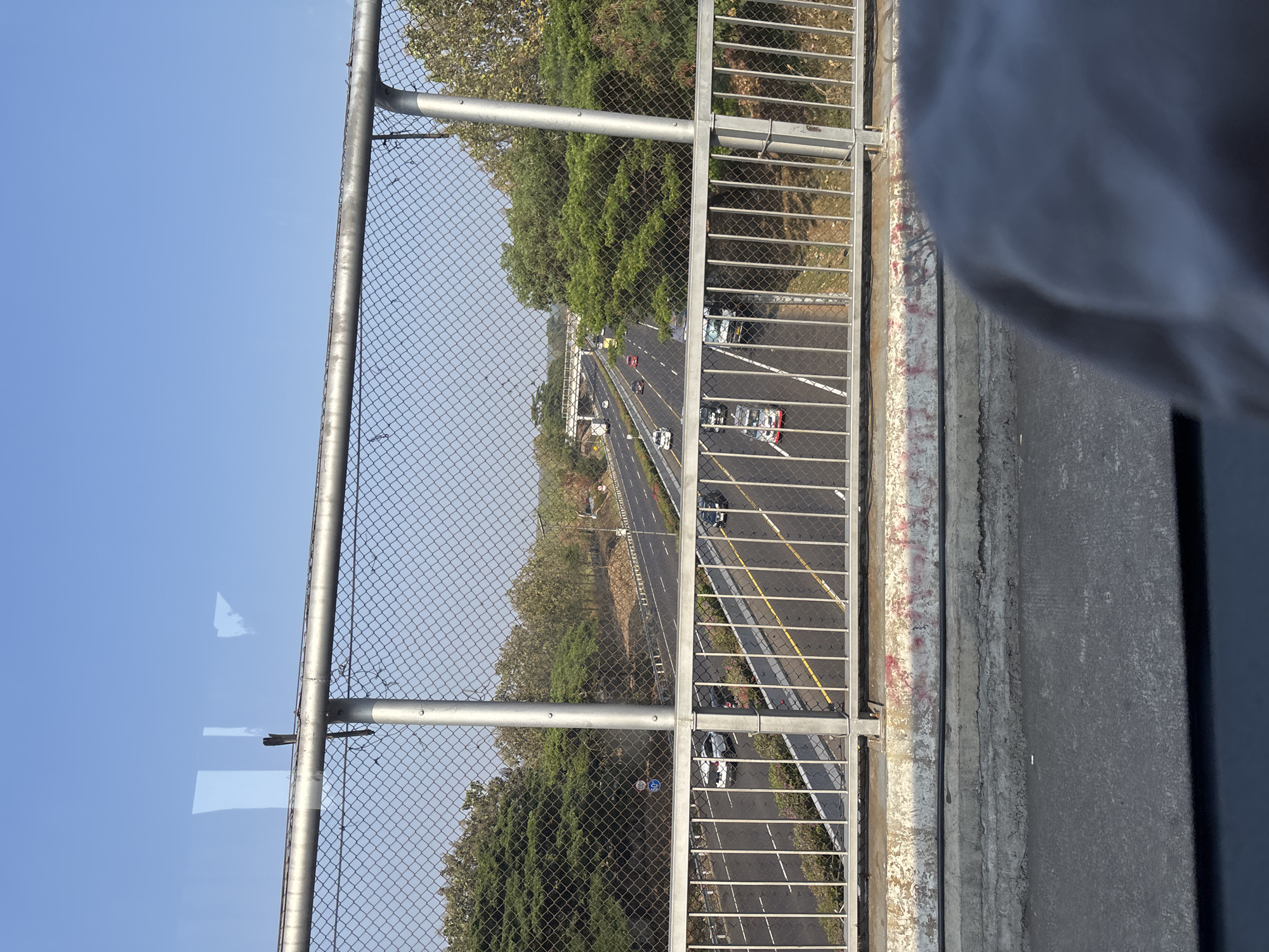The image shows an overhead view of a highway in Cikampek, Indonesia, as seen from a pedestrian overpass. The highway is multi-lane and appears to be busy with traffic, including cars, trucks, and motorcycles. The surrounding environment consists of trees and vegetation, suggesting a semi-rural or suburban setting. The overpass has a chain-link fence for safety. The sky is clear and blue, indicating good weather. The perspective suggests the photo was taken from a moving vehicle or a position slightly elevated above the highway. The foreground shows the concrete structure of the overpass and a portion of what appears to be the roof of a vehicle.