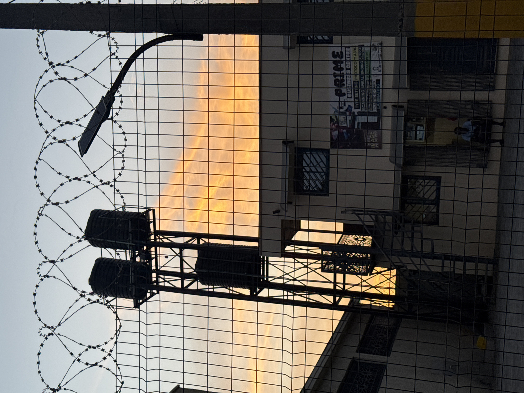The image is a low-angle shot taken through a wire fence with barbed wire at the top. A street lamp is visible on the left side, secured to the fence. Behind the fence is a building with multiple windows, a metal staircase, and water tanks on a metal frame on the roof. There's a sign on the building that reads "PRIME BUSINESS CENTRE." The sky behind the building shows a sunset with orange and yellow hues. A person in white and dark blue uniform can be seen sitting in the shade outside a room. The overall setting appears to be urban or commercial, possibly in Ado, Nigeria.