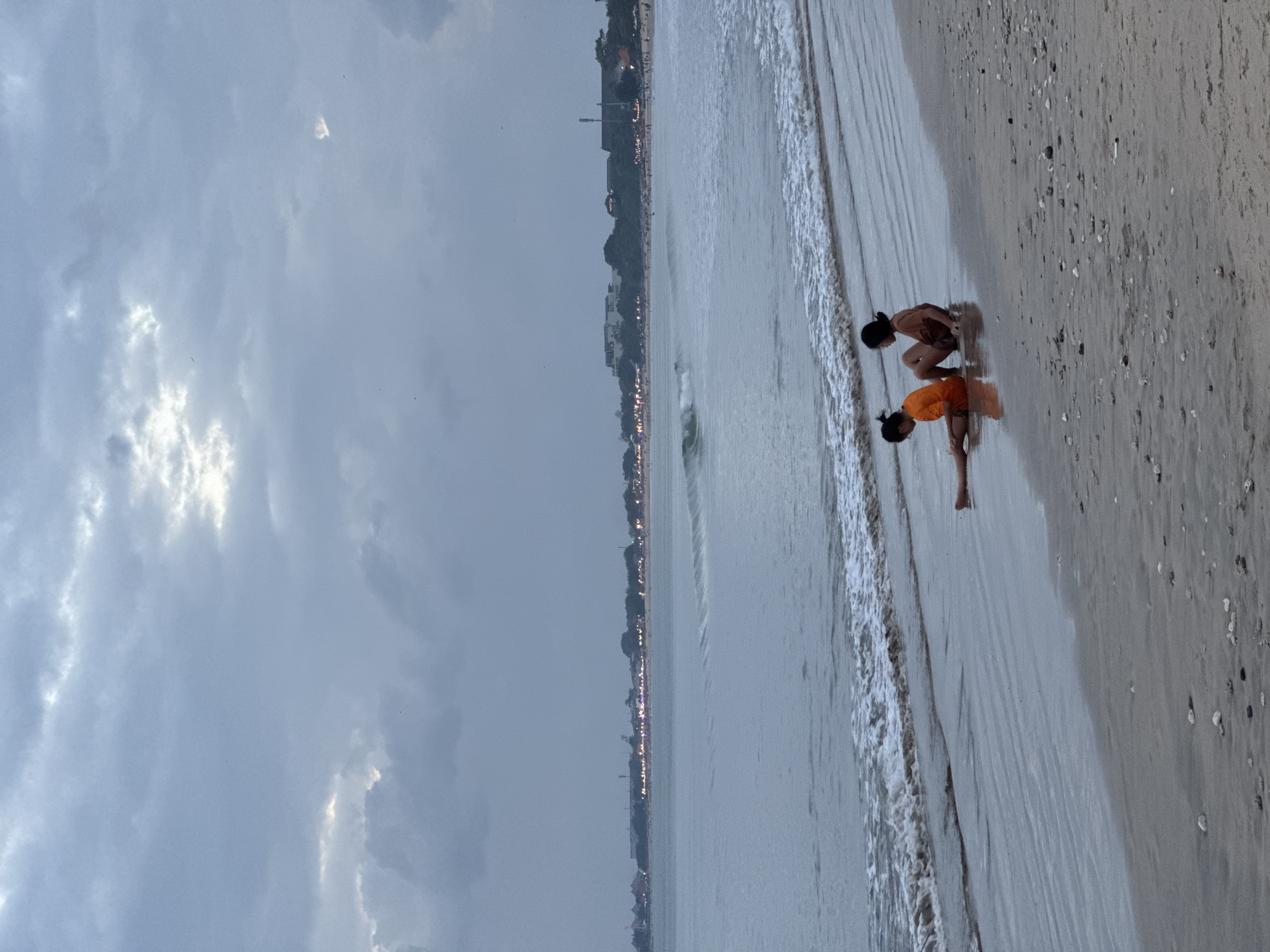 Two young girls are playing on a sandy beach near the water's edge, with the ocean gently washing ashore. One girl, wearing a brown top and shorts, is crouching low, her hands near the sand. The other girl, in an orange top, is also crouched, facing away from the viewer. The scene is set during what appears to be dusk or an overcast day, with soft, diffused light illuminating the clouds. In the background, a distant coastline is visible, dotted with buildings and illuminated by numerous lights, suggesting an inhabited area. The sea is calm with small waves, and the sand is wet and reflective near the waterline, sprinkled with small shells. The overall atmosphere is tranquil and suggests a quiet moment of childhood play. No visible text is present.