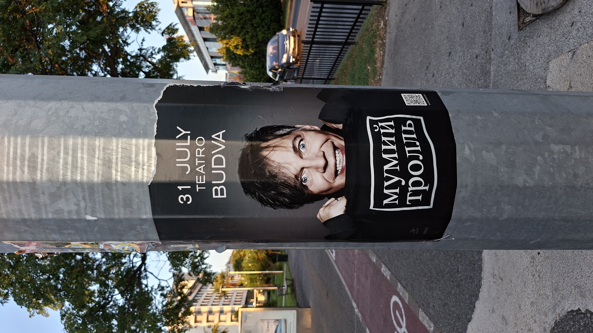 The image shows a poster attached to a metal pole in an urban environment. The poster advertises an event with the text "31 JULY TEATRO BUDVA" and features a close-up of a man's face with wide-open eyes and a smile. Below the man's image, the text "МУМИЙ ТРОЛЛЬ" is displayed in a white box. A QR code is also visible on the poster.
The pole is positioned on a sidewalk next to a street where a car is visible in the background. The street appears to be a road with a metal fence alongside it. Trees and buildings are present in the background, suggesting a city setting. The sidewalk has a red-patterned surface, possibly indicating a bike lane or pedestrian walkway. The lighting suggests it is daytime, with shadows indicating sunlight.
The location is Podgorica, Montenegro, as indicated by the poster's text referring to "Teatro Budva", a cultural venue in Budva, Montenegro, and the presence of Cyrillic script on the poster, which is used in Montenegro. The overall scene depicts a street in Podgorica with a poster advertising an event.